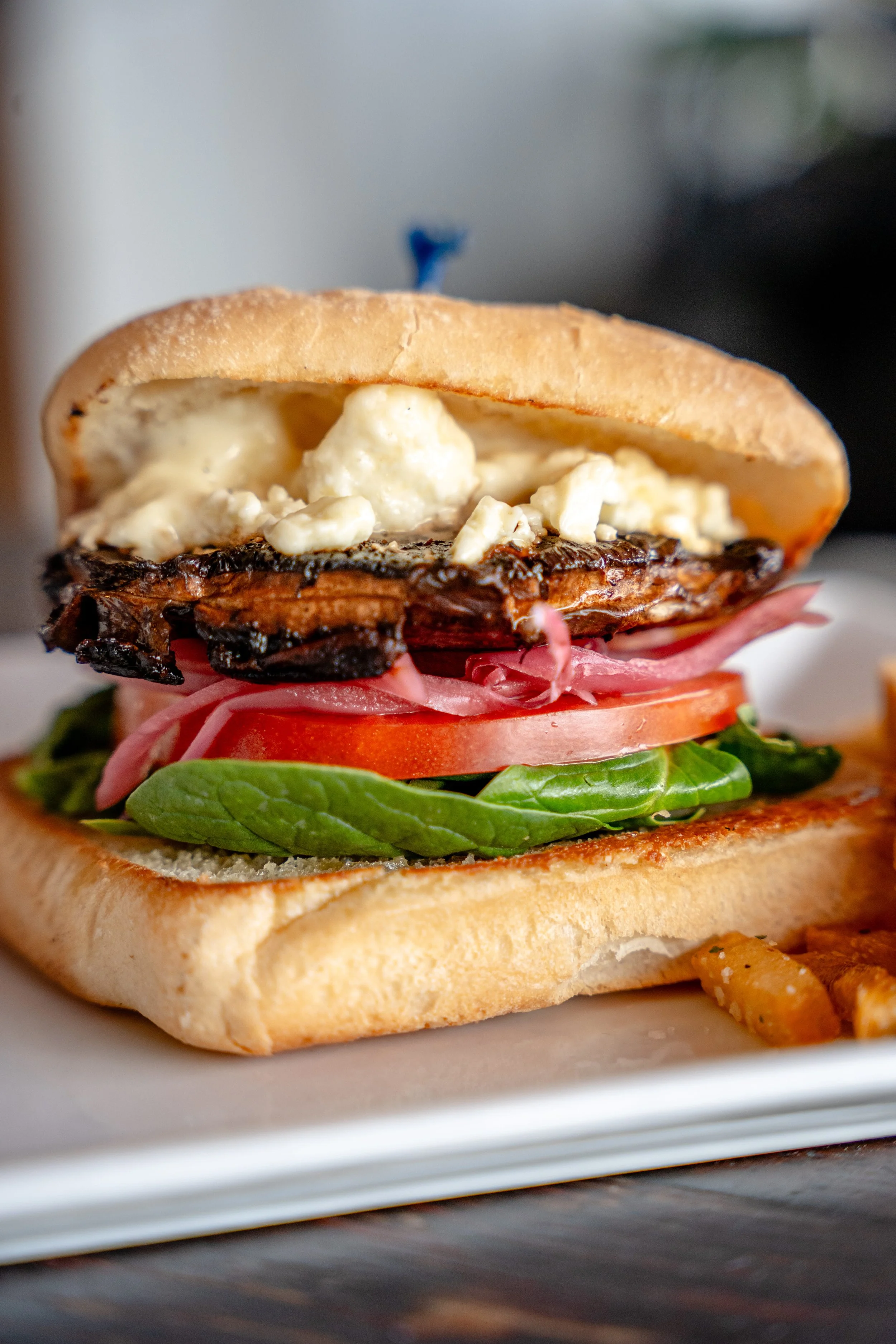 Close-up of a sandwich with lettuce, tomato, sliced ham, grilled eggplant, and potato salad on a bun, with French fries on the side.