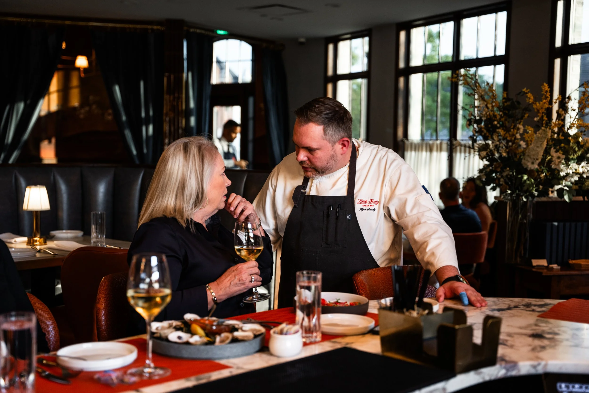 A female customer with blonde hair holding a glass of white wine, talking to a male chef wearing a white chef's coat and black apron in a restaurant.