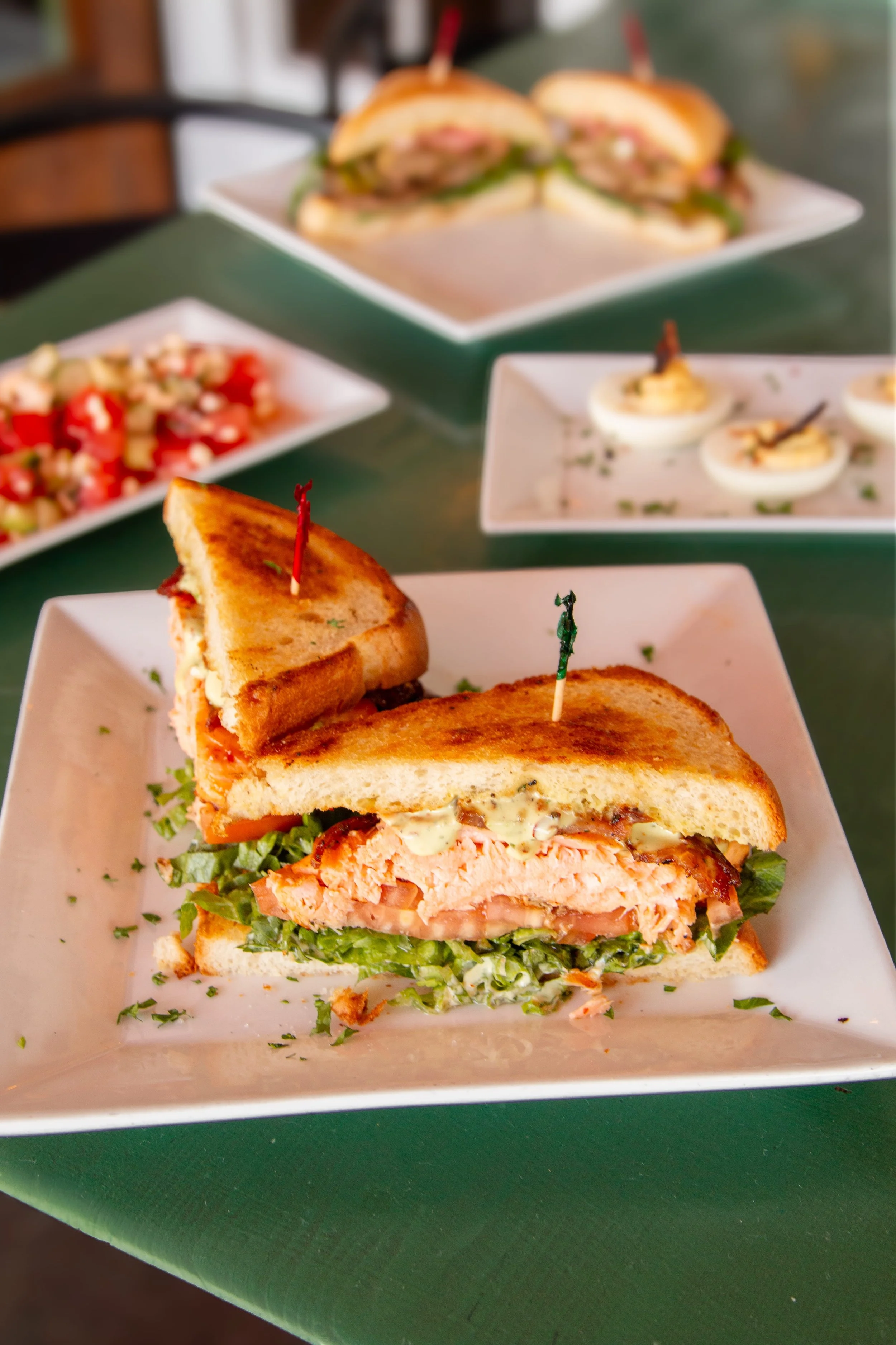 A club sandwich with toasted bread, lettuce, tomato, chicken, and mayonnaise, secured with skewers, served on a square white plate. In the background, various dishes including salads, deviled eggs, and sandwiches on plates are visible.