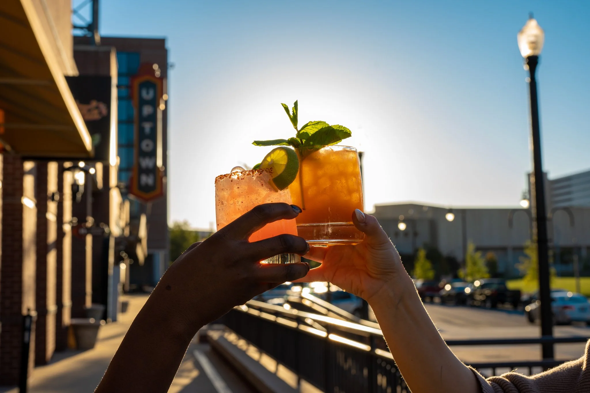 Two hands holding up a glass of orange-colored cocktail with lime and mint garnishes, backlit by the setting sun in an outdoor urban setting.
