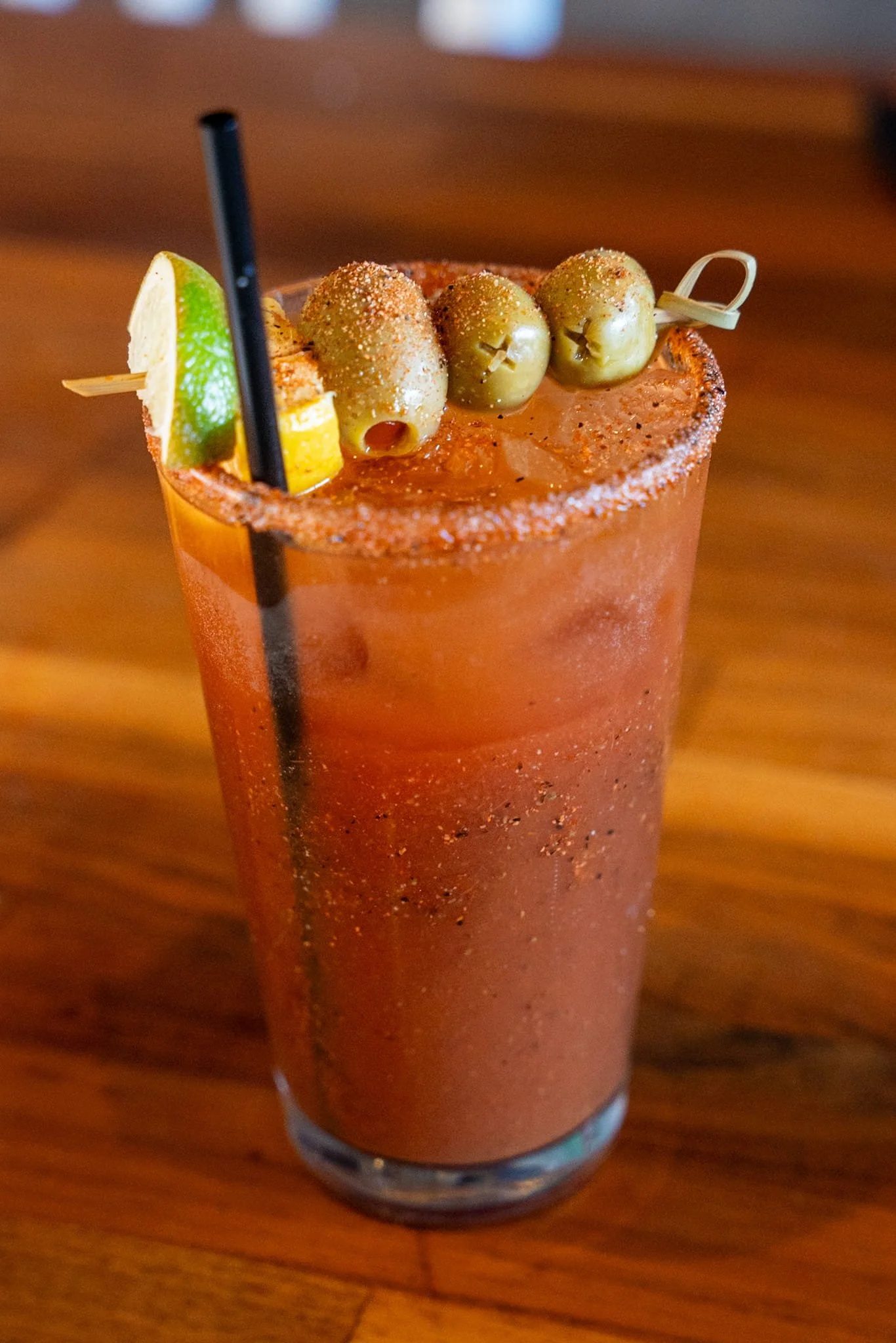 A bloody mary cocktail garnished with green olives, lemon wedge, and a lime wedge, with a salted rim and two black straws, on a wooden table.