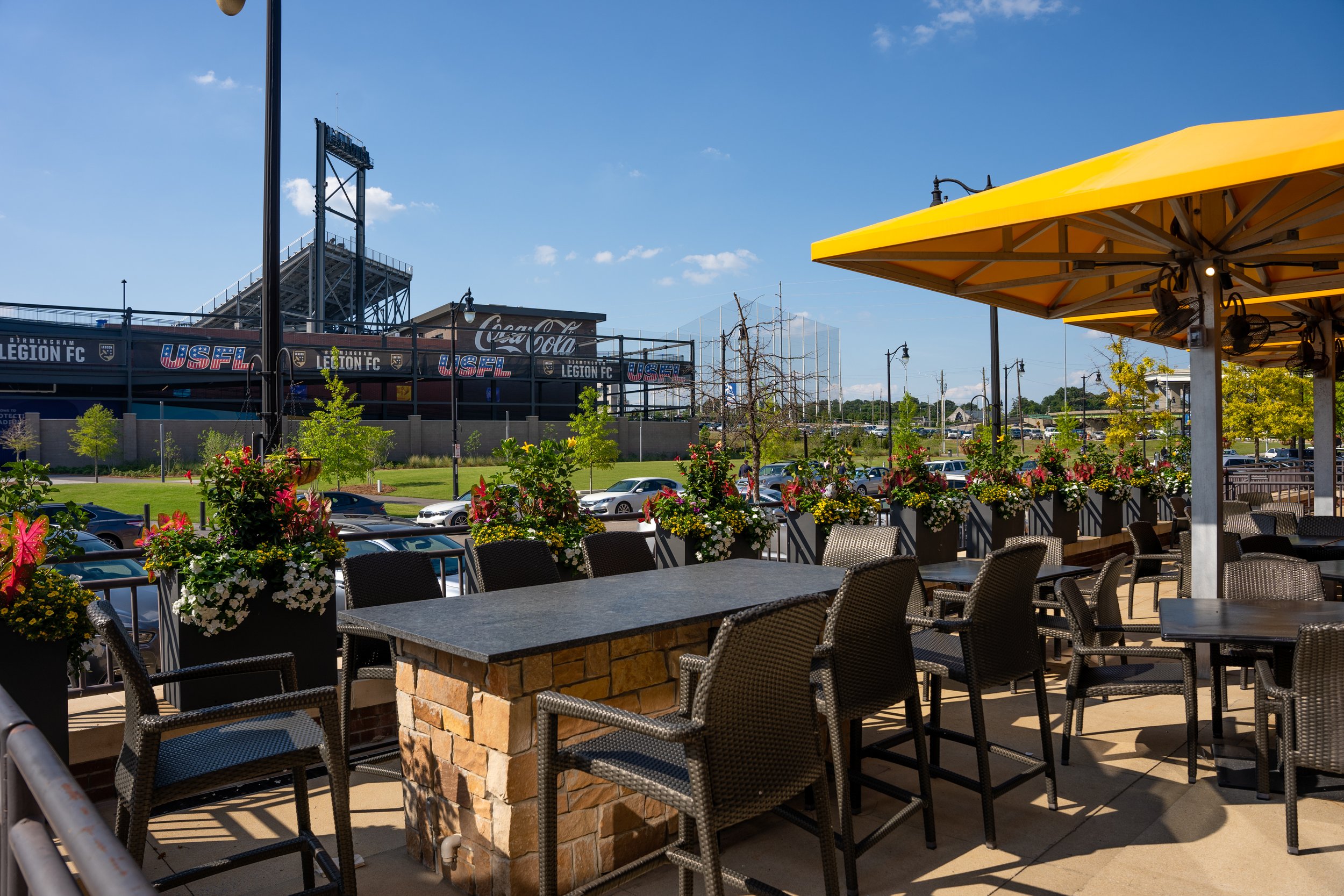 Outdoor seating area at a restaurant or cafe with tables, chairs, and yellow umbrellas, overlooking a parking lot and a stadium in the background under a blue sky.
