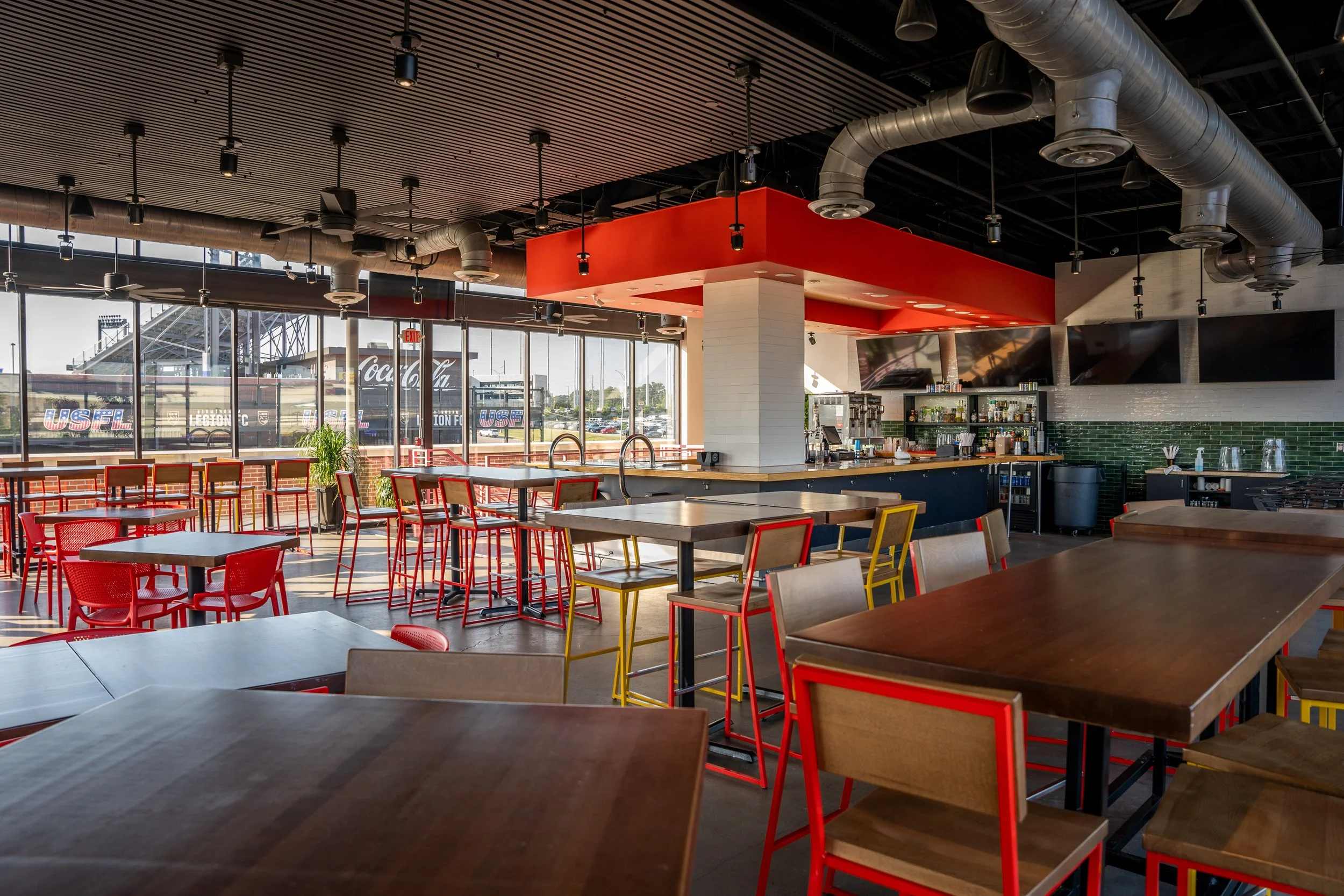 Interior of a modern, brightly lit bar or restaurant with red, yellow, and wood chairs around wooden tables, a bar area with beverages and equipment, and large windows showing a sports stadium outside.