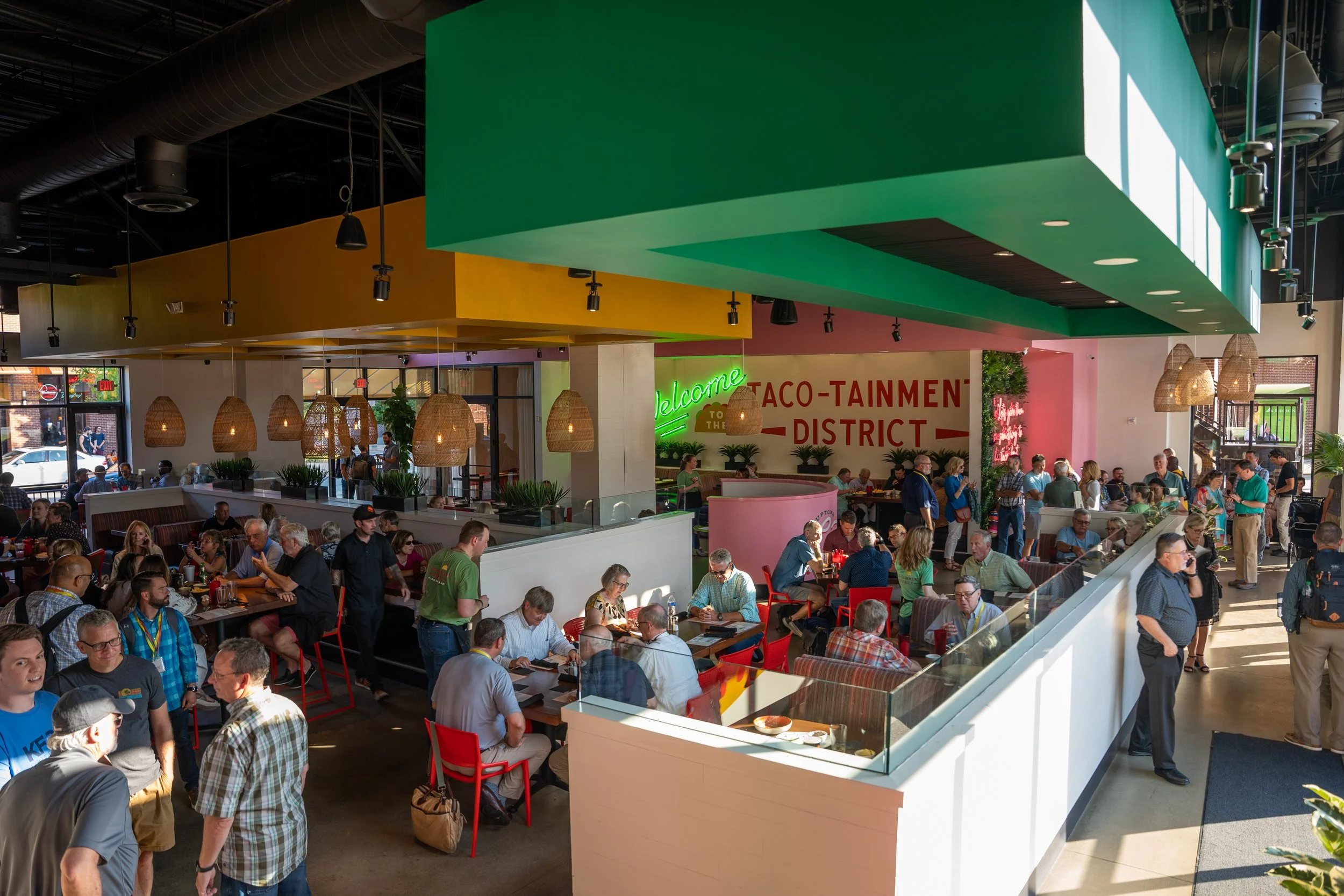 Crowded restaurant with colorful decor, including green, yellow, and pink ceiling sections, and a neon sign that says "Welcome". People are seated at tables and standing in line, enjoying their meals in a lively atmosphere.