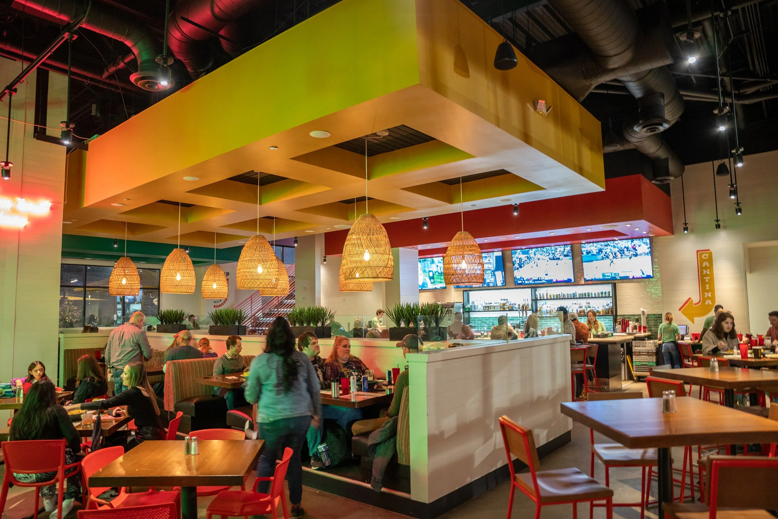 Interior of a bustling restaurant with colorful ceiling and hanging wicker lights; patrons seated at tables and a bar in the background, with a sign pointing to the cantina.