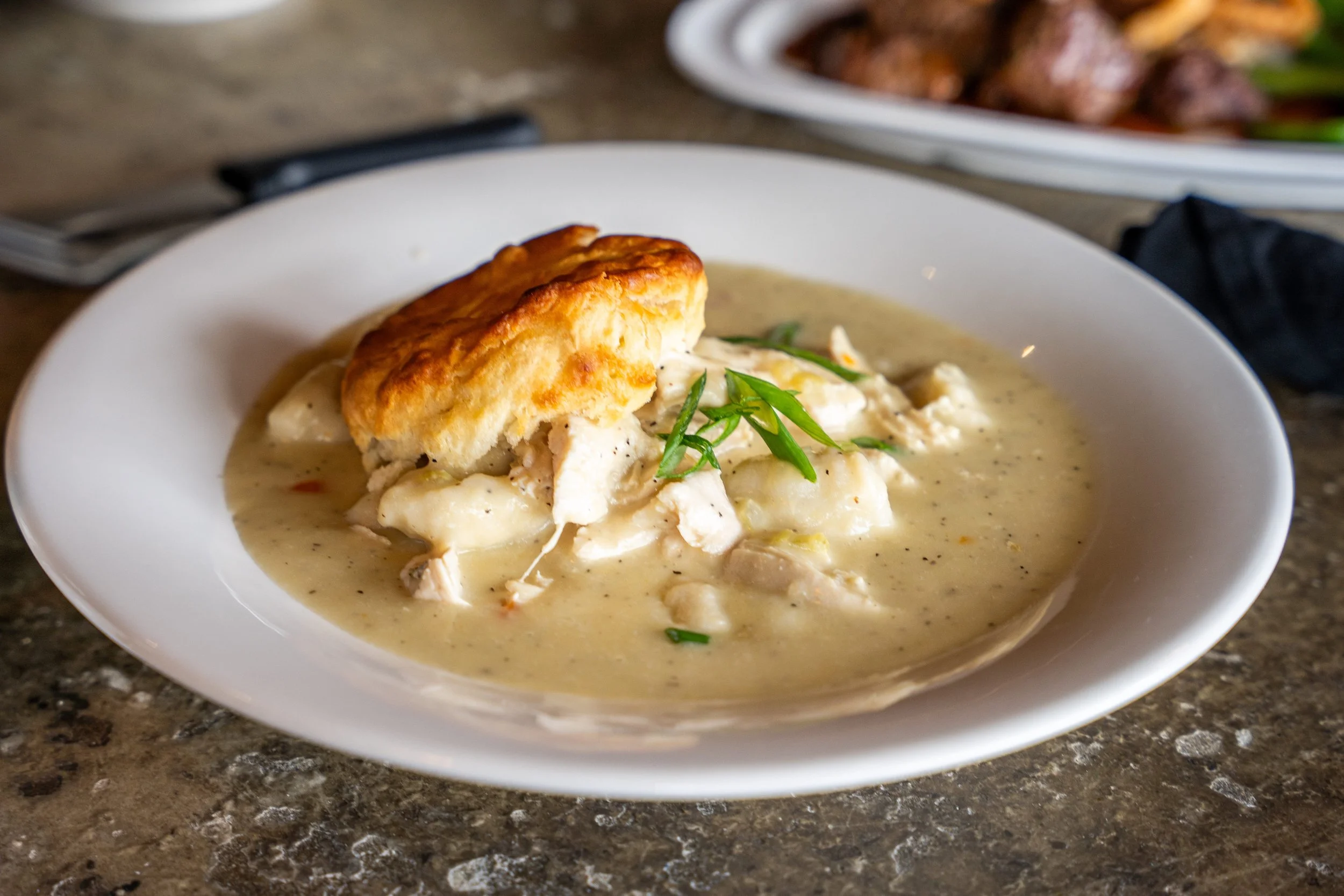 A white bowl containing chicken pot pie with a flaky crust, topped with green onions, on a textured countertop.
