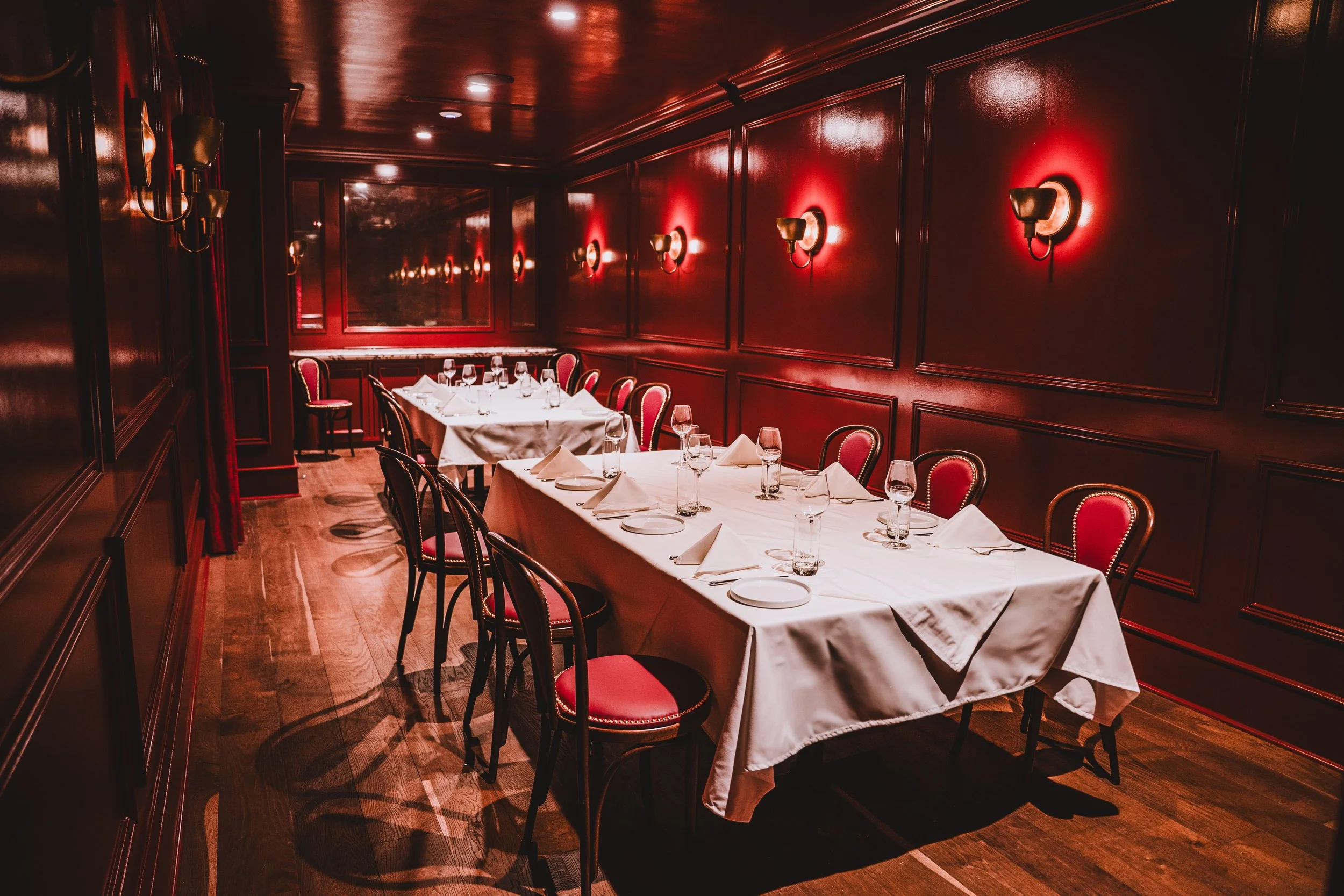 Elegant restaurant dining room with a long table set with white tablecloths, napkins, wine glasses, and plates, surrounded by red velvet chairs, with red-paneled walls and wall-mounted lights.