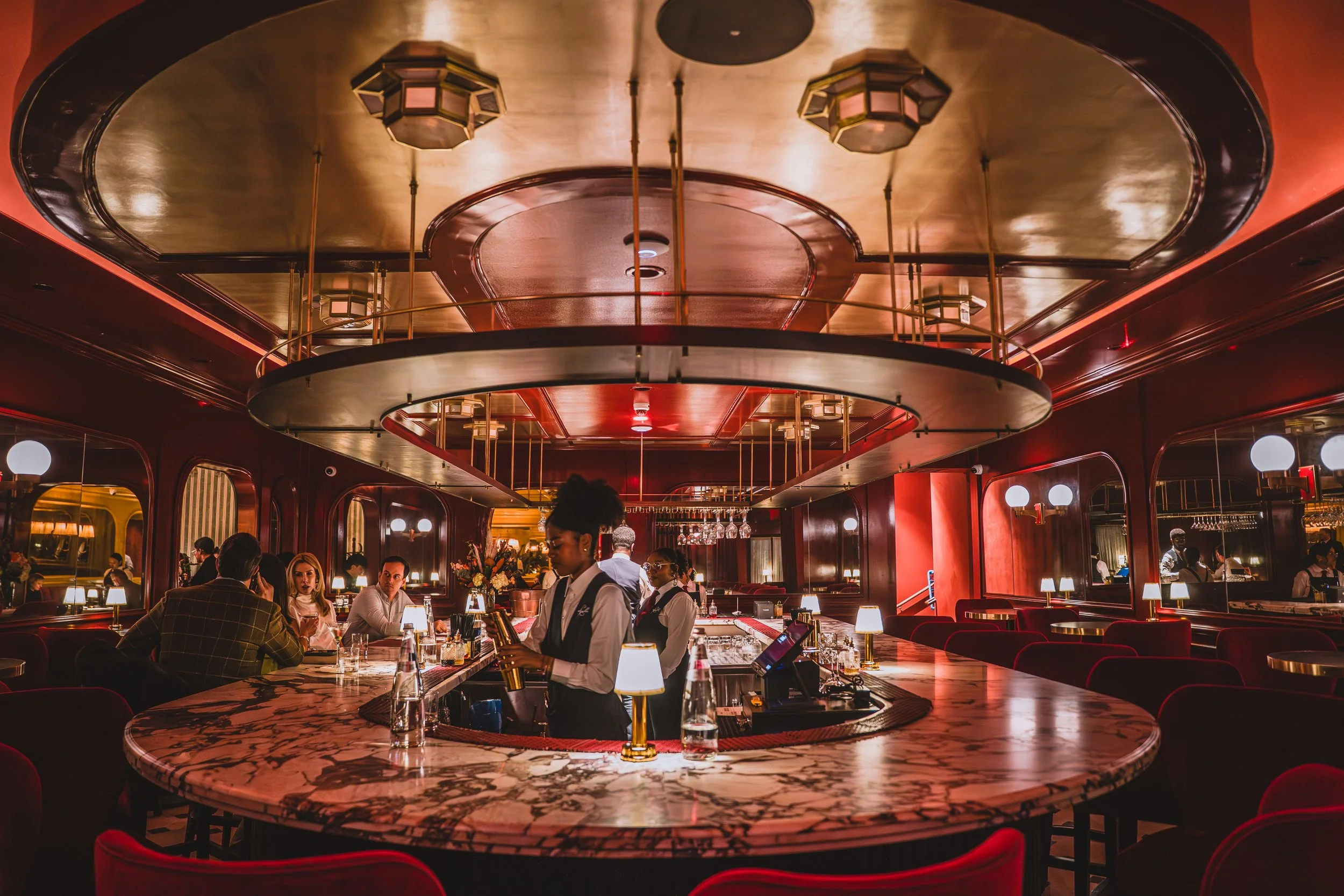 Interior of an elegant restaurant or bar with red walls, reflective mirrors, and a marble-topped bar counter. There are people dining and bartenders preparing drinks, with ambient warm lighting and modern decor.