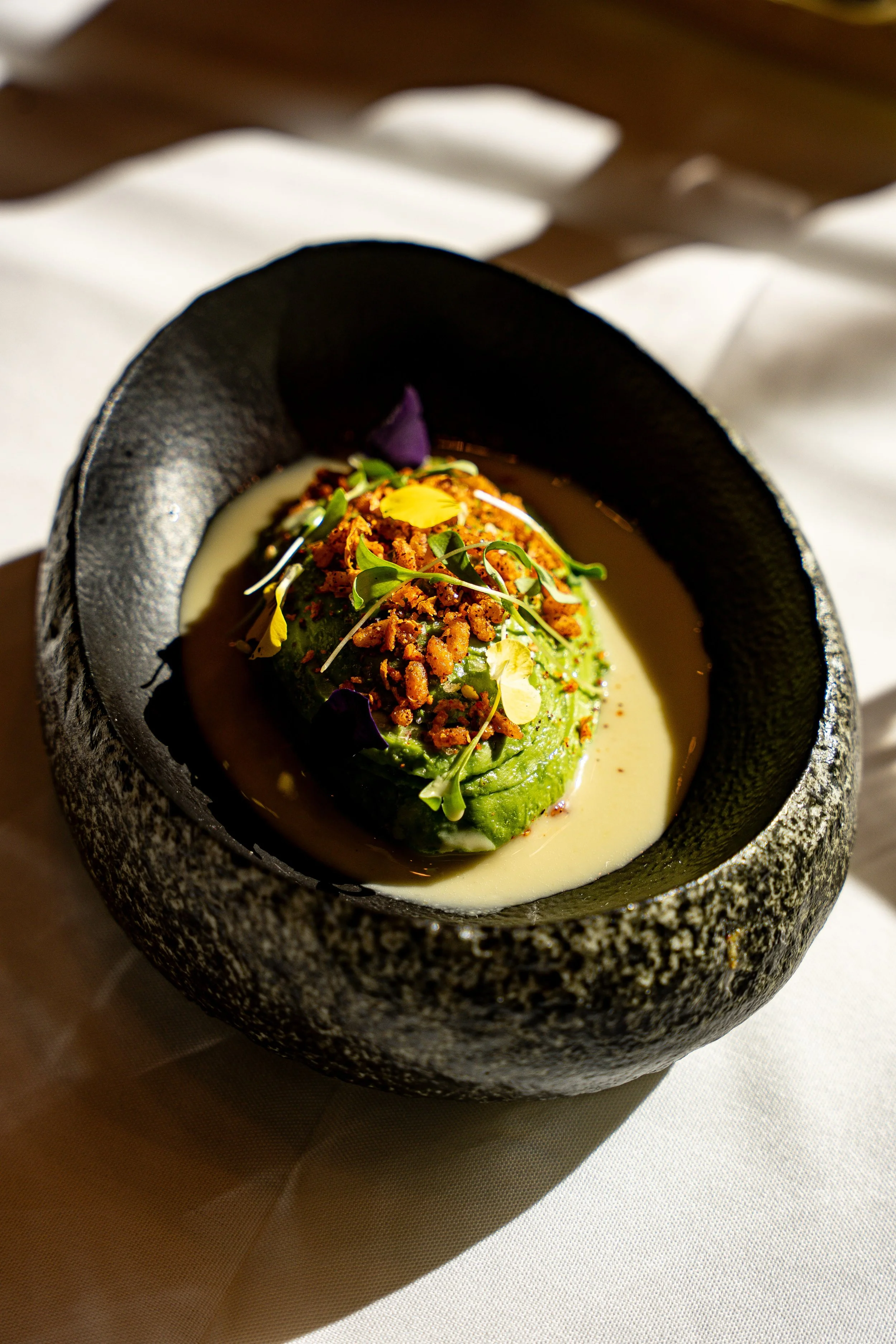 A gourmet dish with a green fill wrapped in avocado, topped with microgreens and crunchy garnishes, served in a black stone bowl with a white sauce or broth.