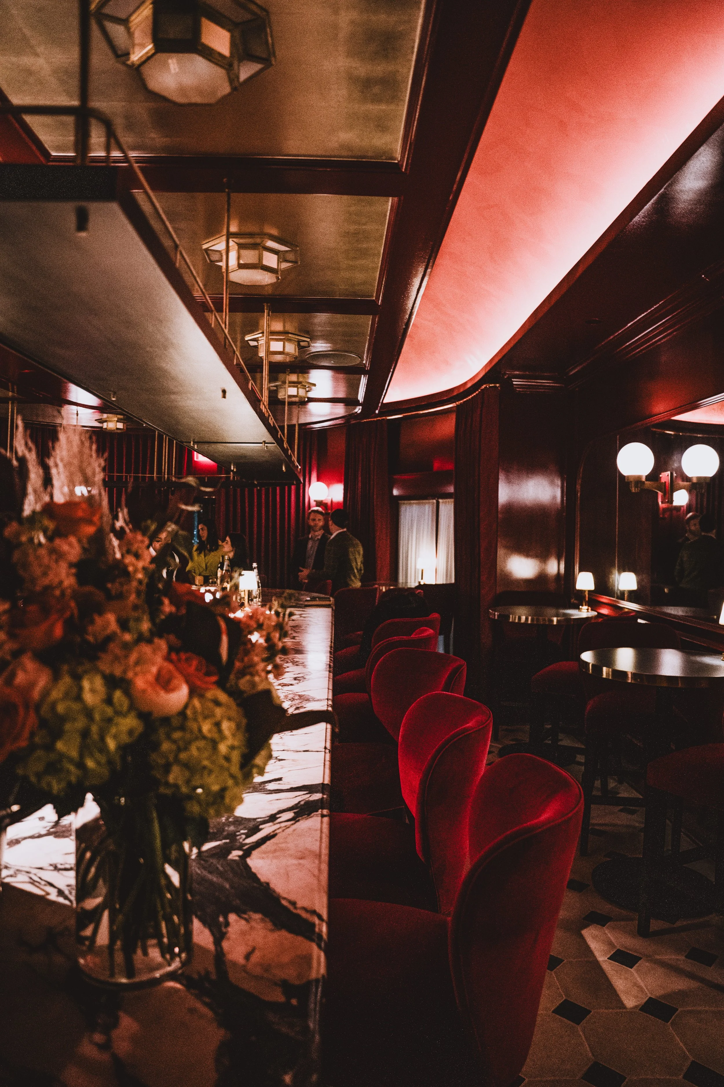 A dimly lit bar or lounge with red velvet chairs, a marble countertop, floral arrangements, and small table lamps, with people standing and conversing in the background.