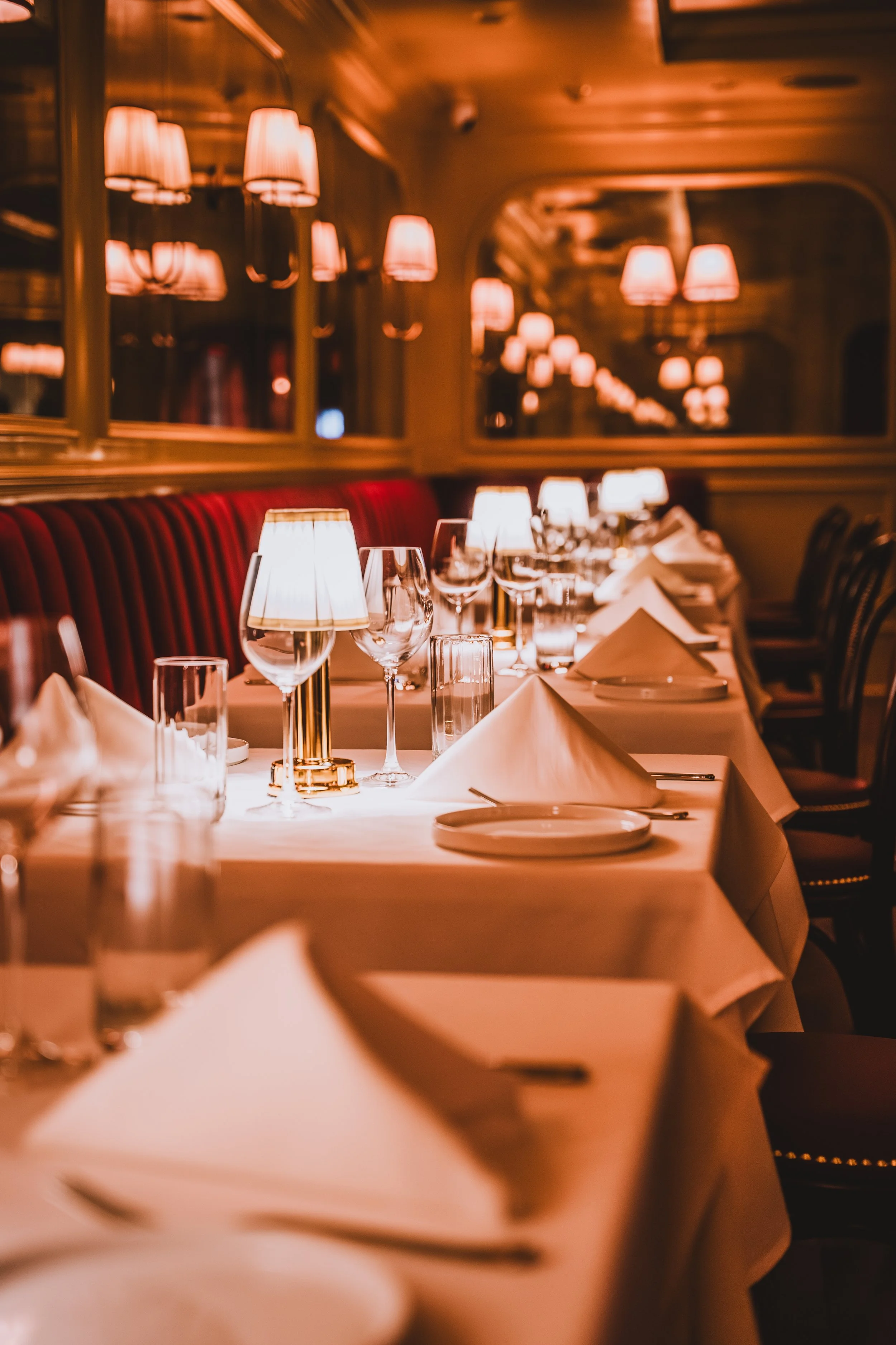 Elegant restaurant dining table set with white tablecloths, folded napkins, wine glasses, water glasses, and small lamps with warm lighting, in a room with dark chairs and mirror wall reflections.