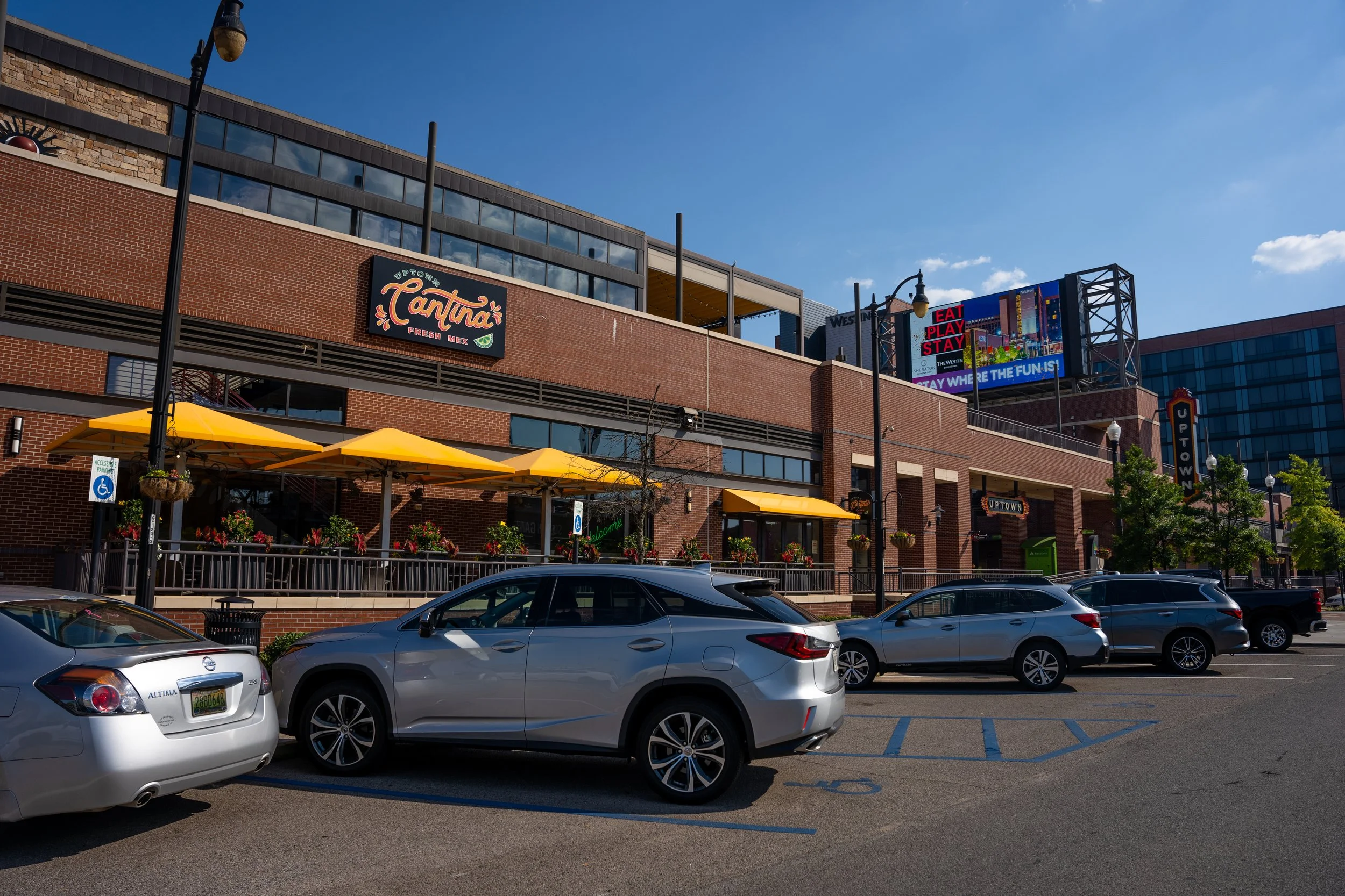 Parking lot in front of a brick building with yellow umbrellas and potted plants, signs for Cantina and Uptown, and a digital billboard displaying "Eat Play Stay" and "Stay Where the Fun Is!" under a blue sky.