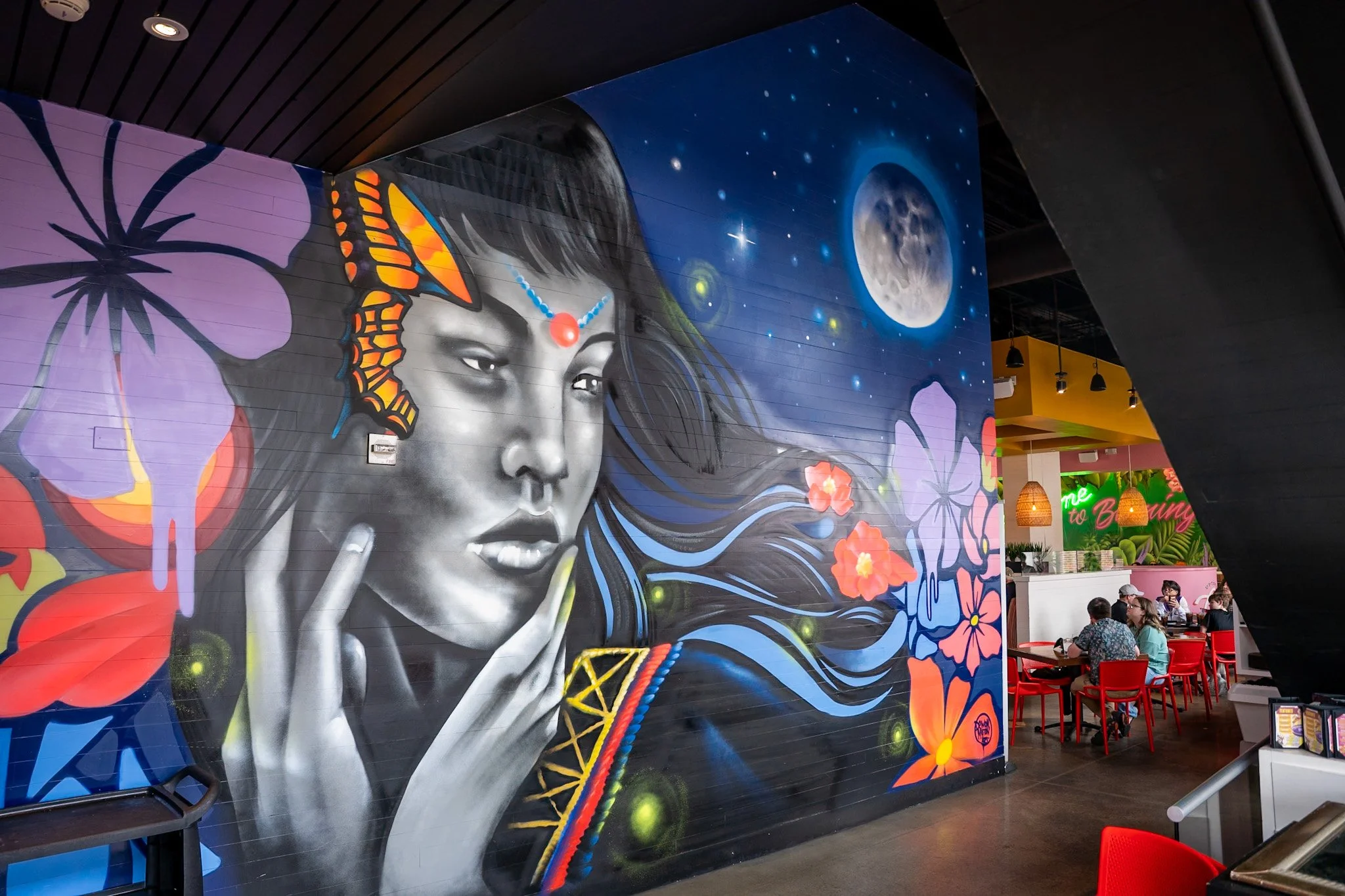 Colorful mural of a woman with long flowing hair, flowers, a butterfly, and a moon in the background, inside a modern restaurant with red chairs and diners.