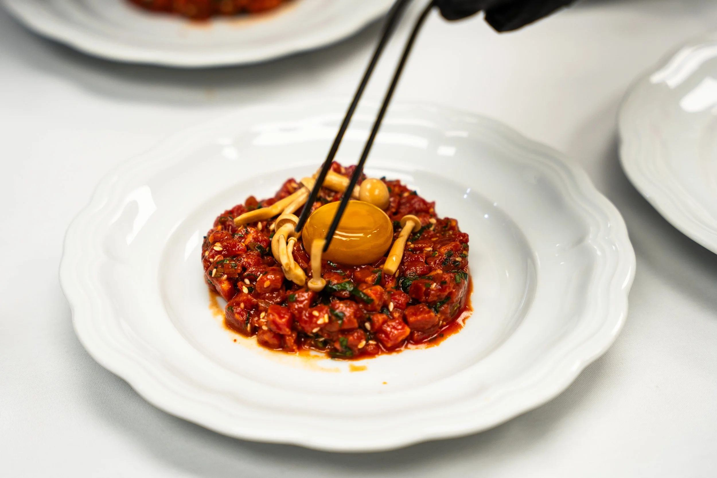 A white plate with a serving of spicy red chili topped with an olive and sliced enoki mushrooms, being pickled or added with black tongs.