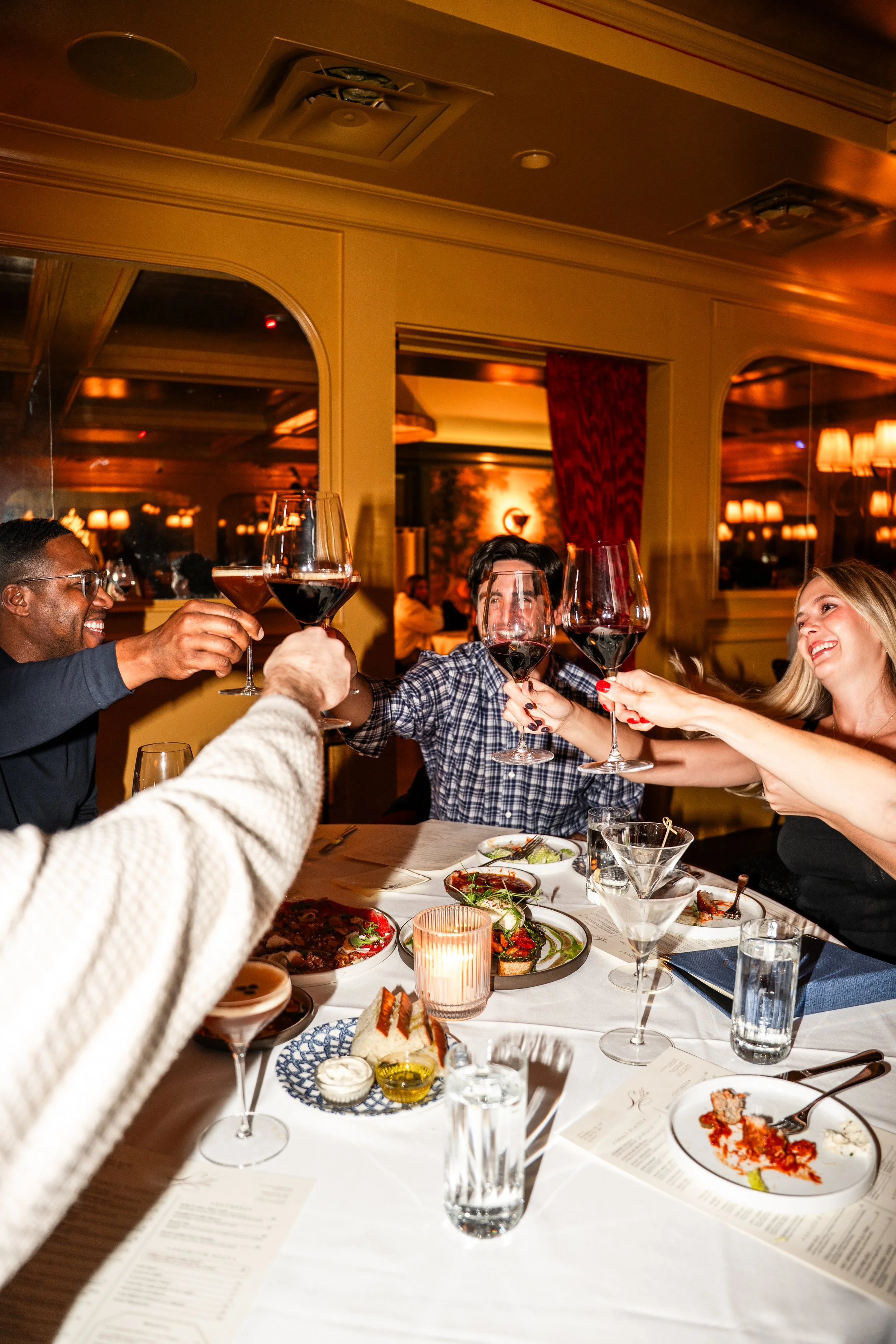 A group of five friends raising glasses of red wine in a toast at a dinner table filled with food and drinks in an elegant restaurant.