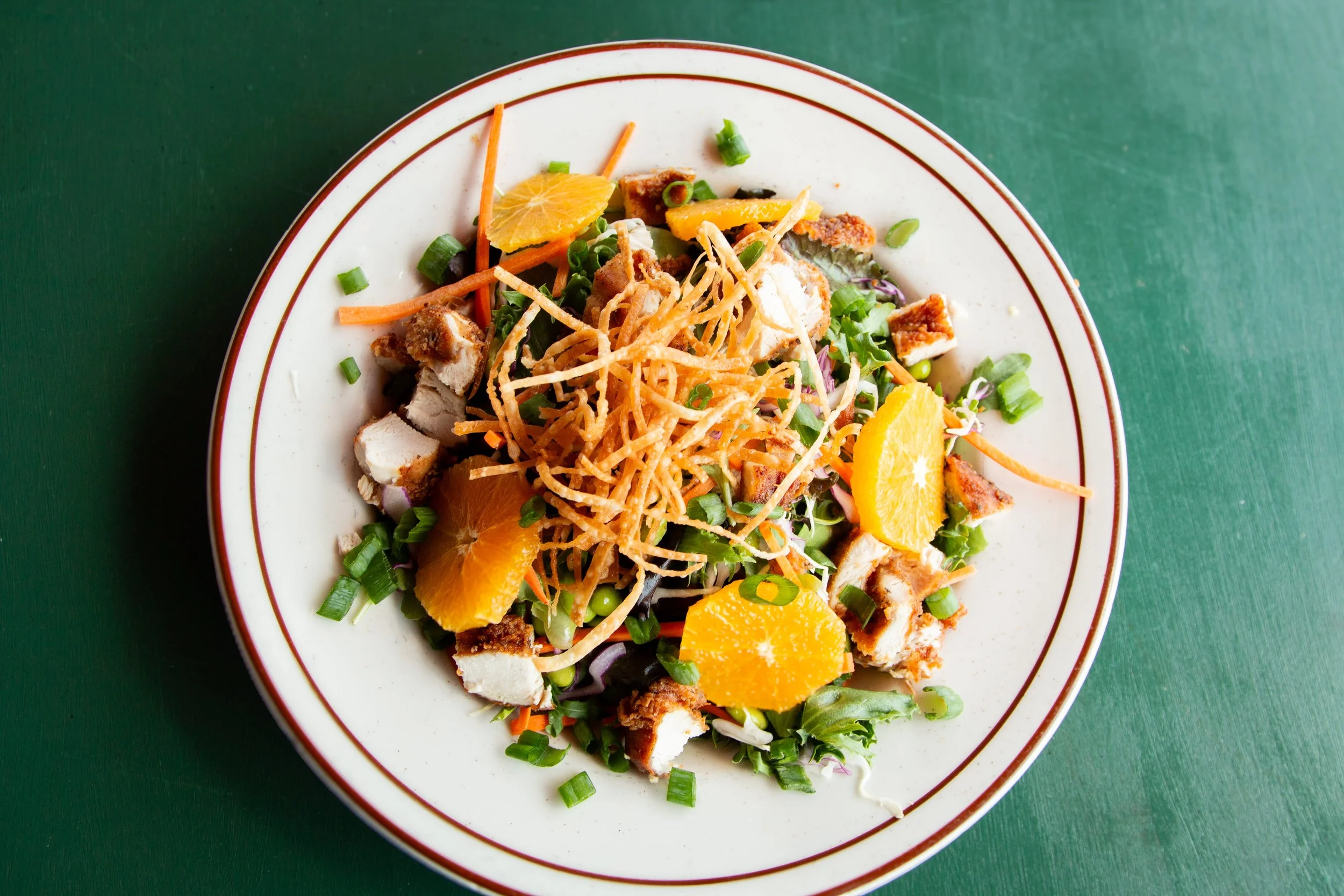 Salad with mixed greens, shredded carrots, orange slices, crispy fried chicken pieces, and crispy noodle strips on a white plate with red trim, set on a green table.