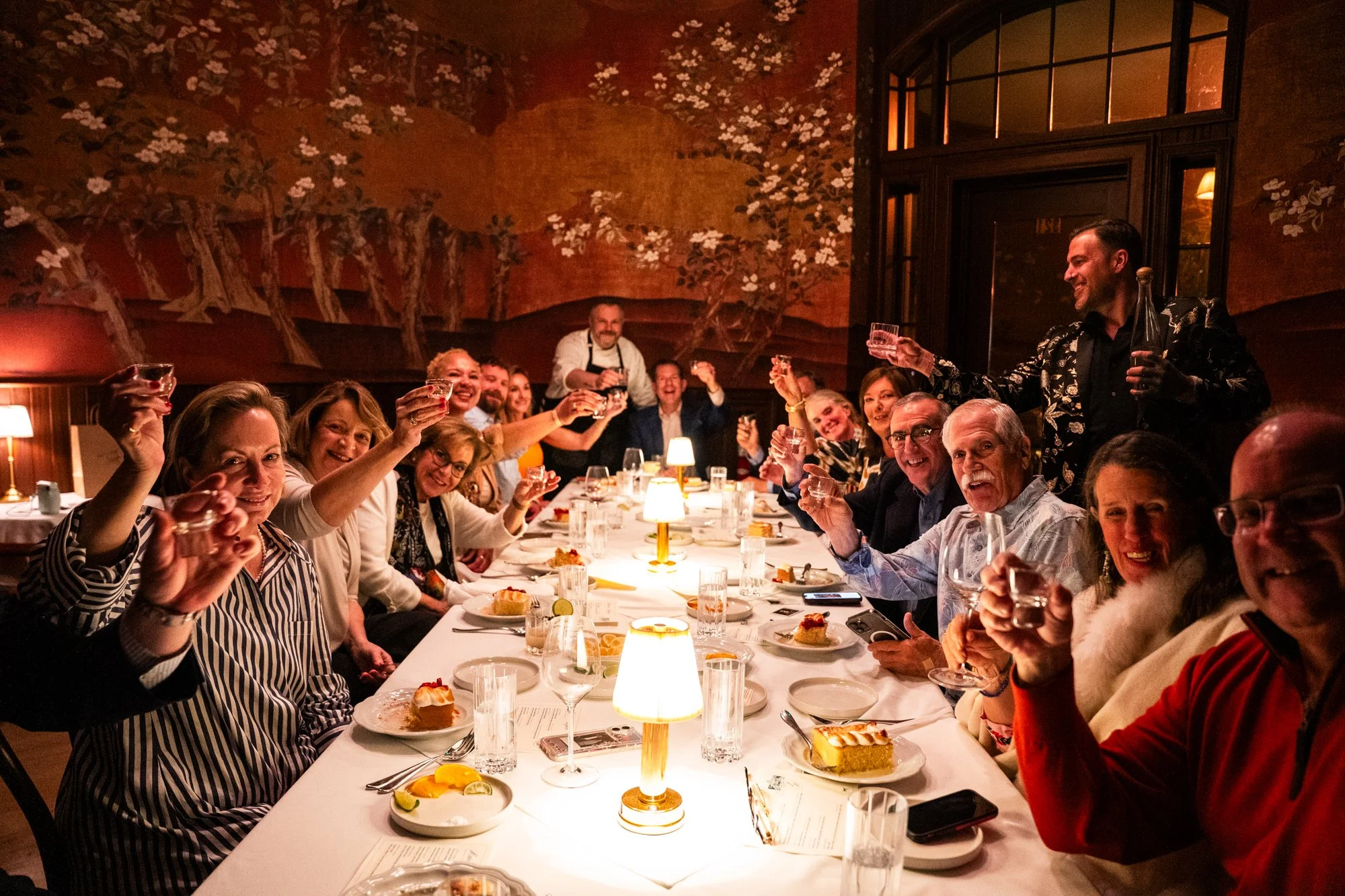 People celebrating at a dinner party, raising glasses in a toast, sitting around a long table with candles and plates of cake in a warmly lit restaurant with a mural background.