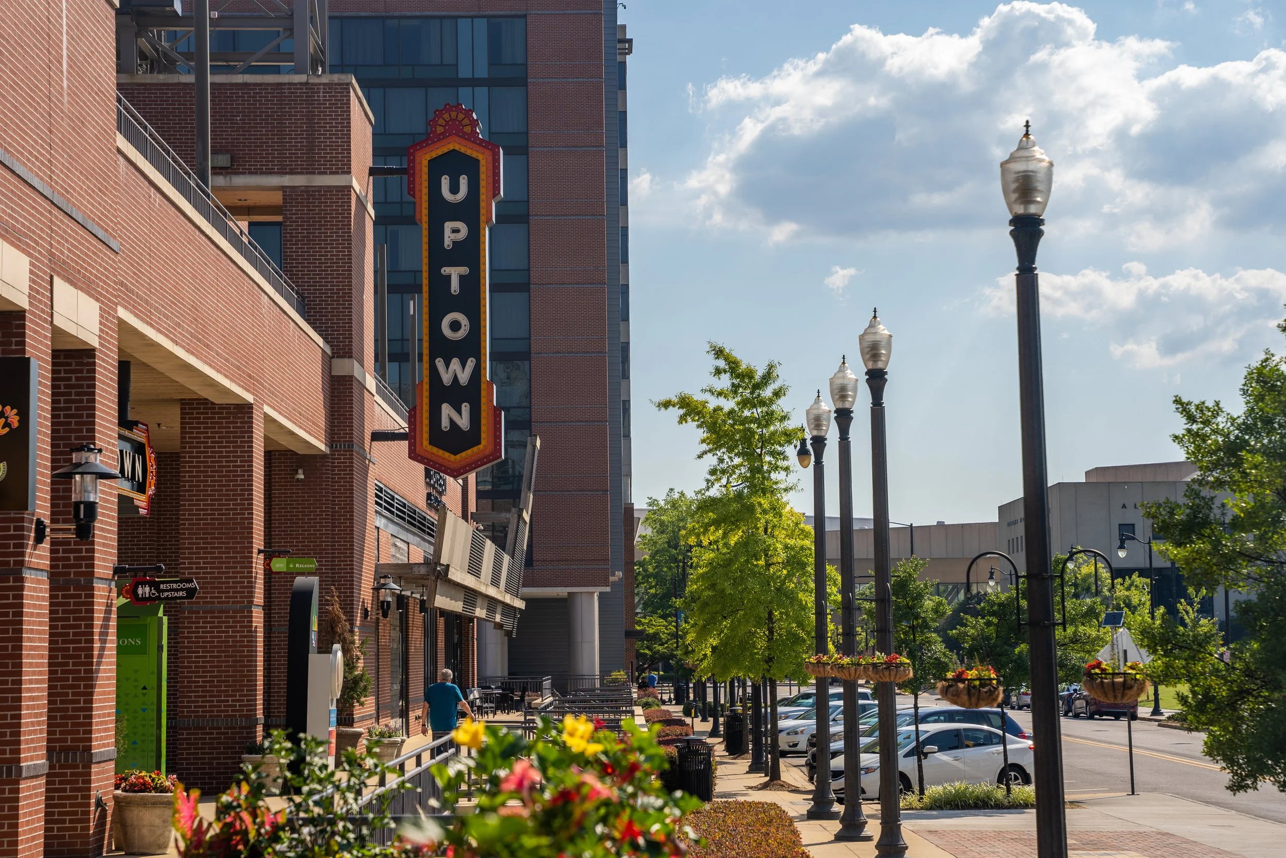 Downtown street view with brick building on left, vintage style lamp posts, trees, and parked cars on the right, under a partly cloudy sky.