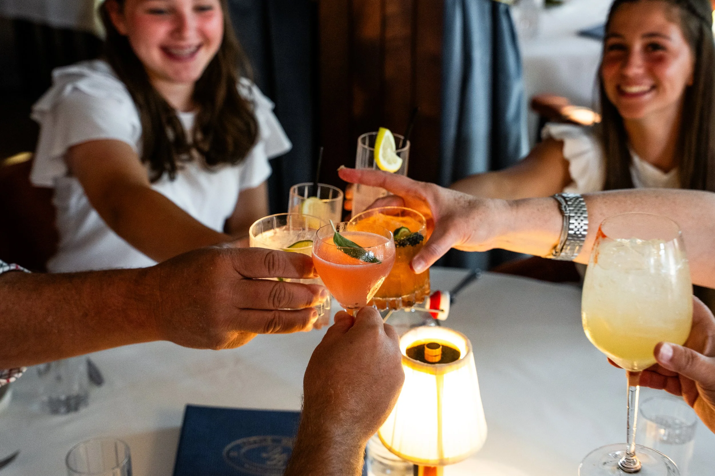 People clinking glasses of cocktails at a table, with smiling women in the background.