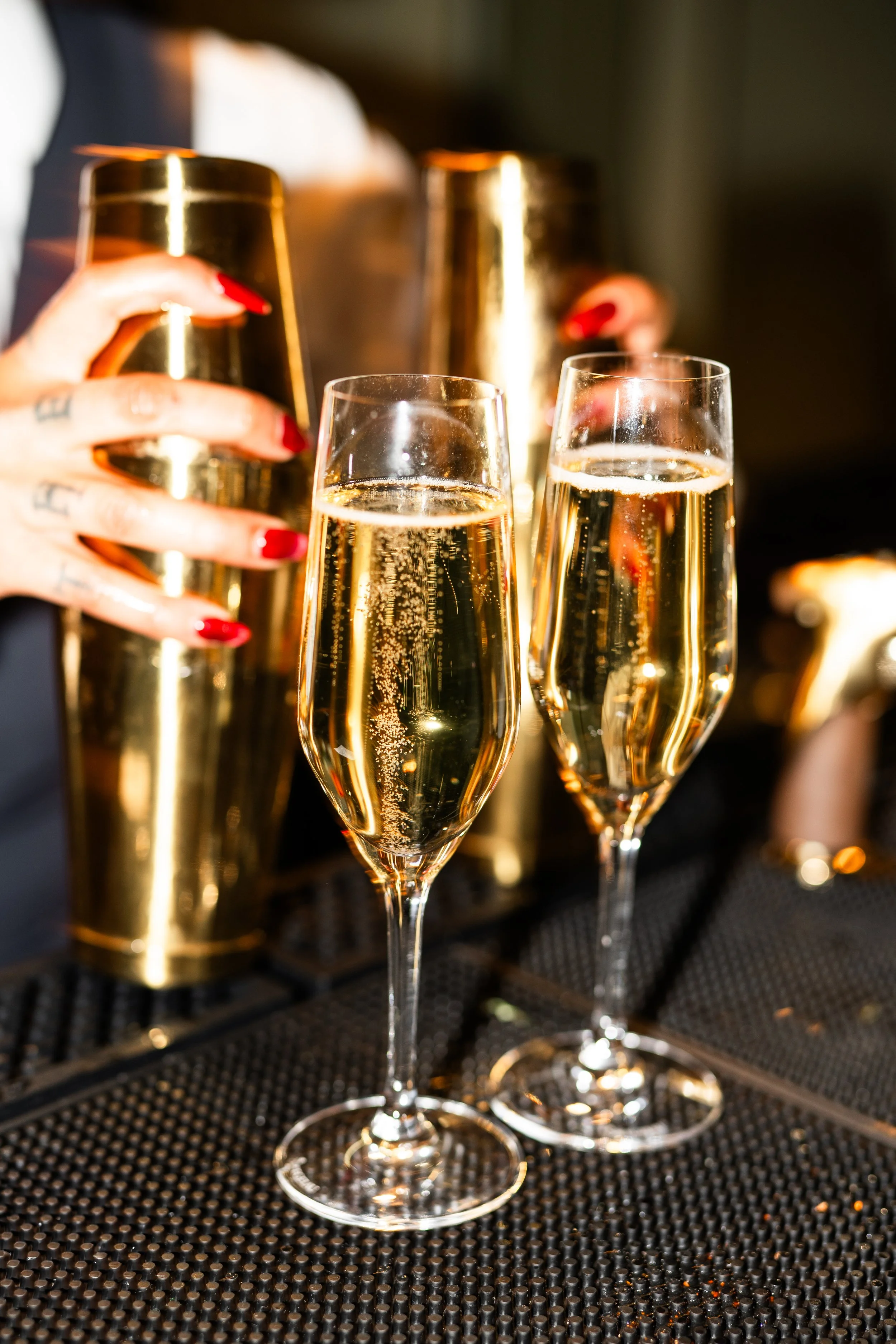 Two glasses of champagne on a bar counter, with a bartender pouring champagne from a gold shaker in the background.