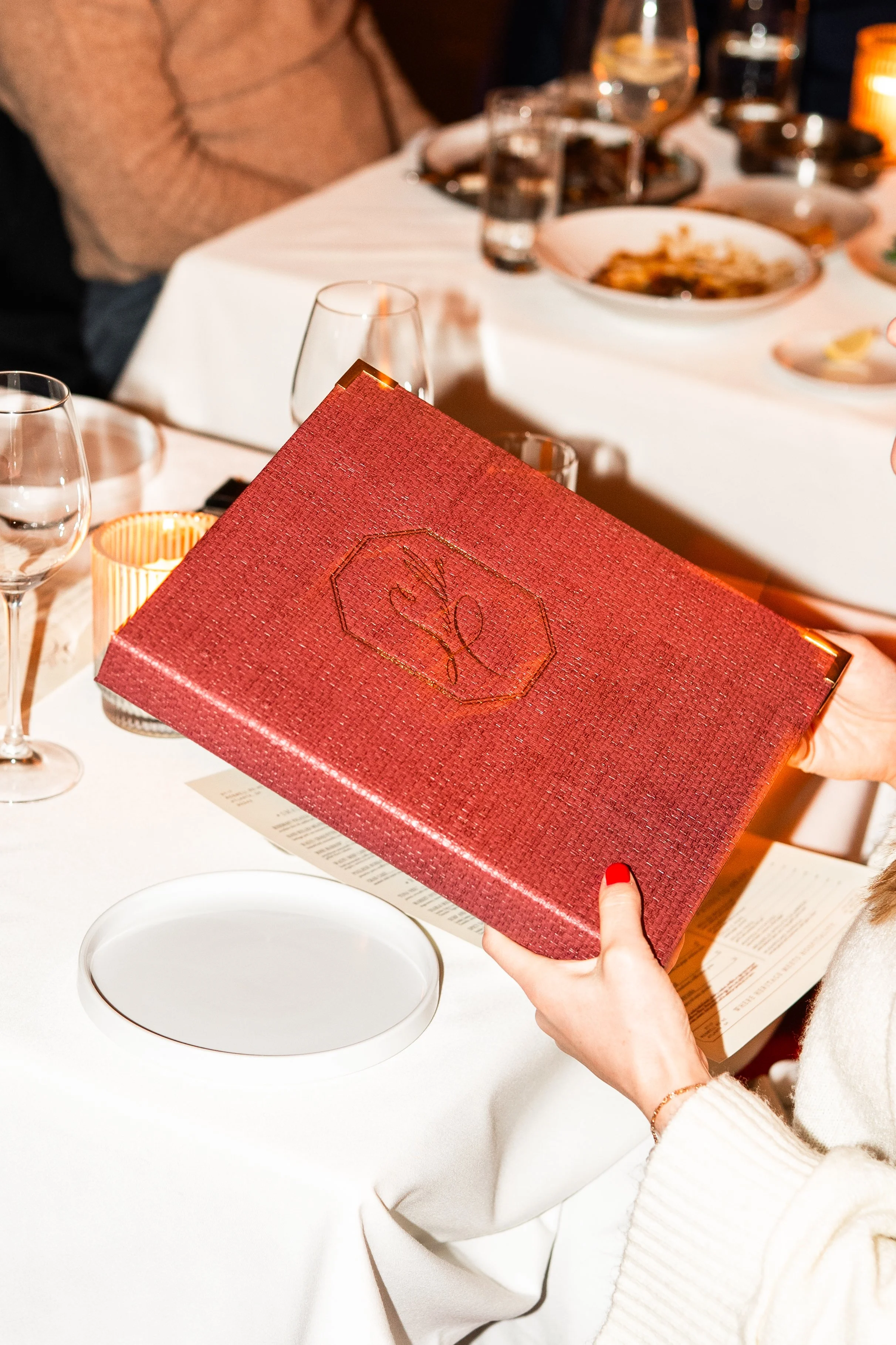 A person holding a red gift box above a dining table with plates, glasses, and cutlery in a restaurant setting.