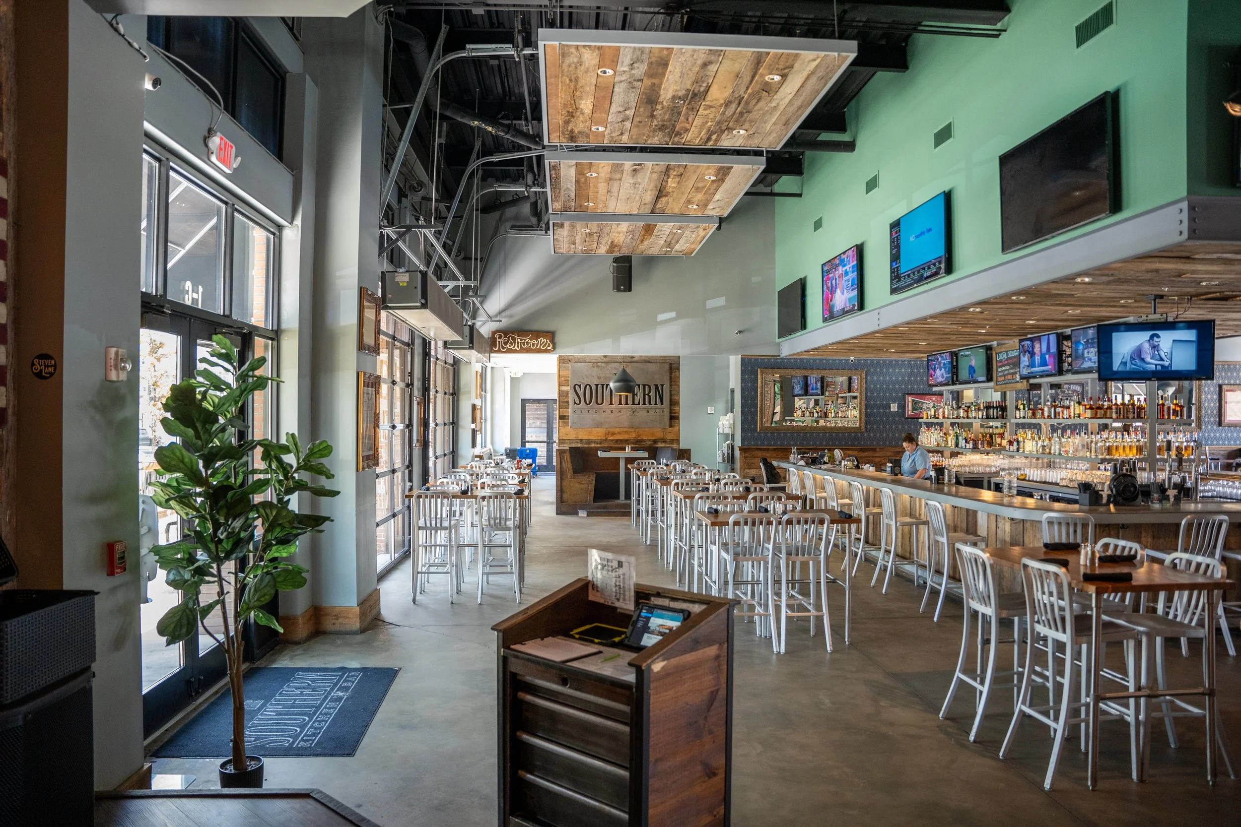 Empty restaurant with bar area and multiple TVs on the wall, barstools around the bar, table seating, potted plant near entrance, and neon sign for restrooms.