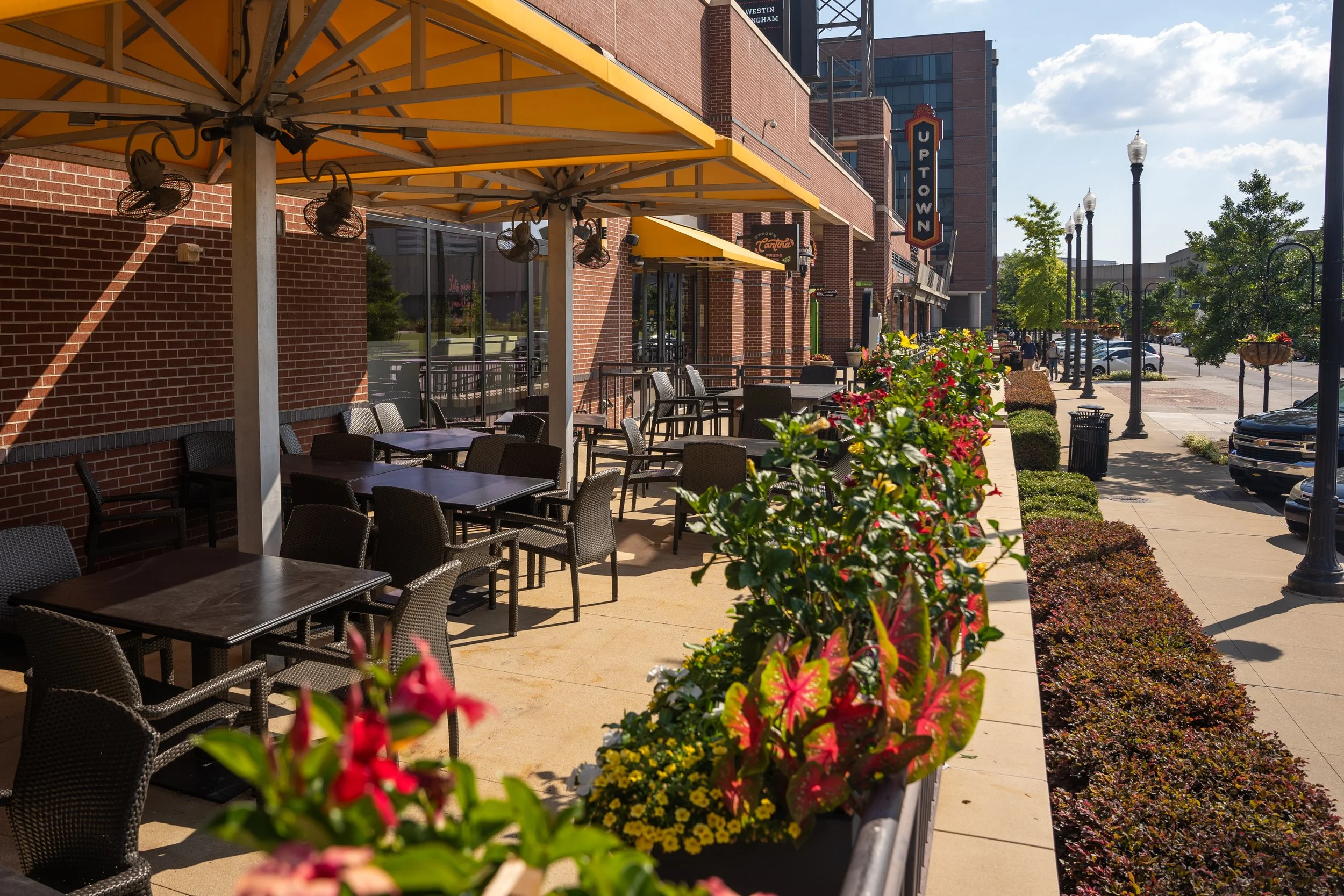 Empty outdoor patio with tables and chairs, yellow umbrellas, and flower boxes along the sidewalk on a sunny day in an urban area.