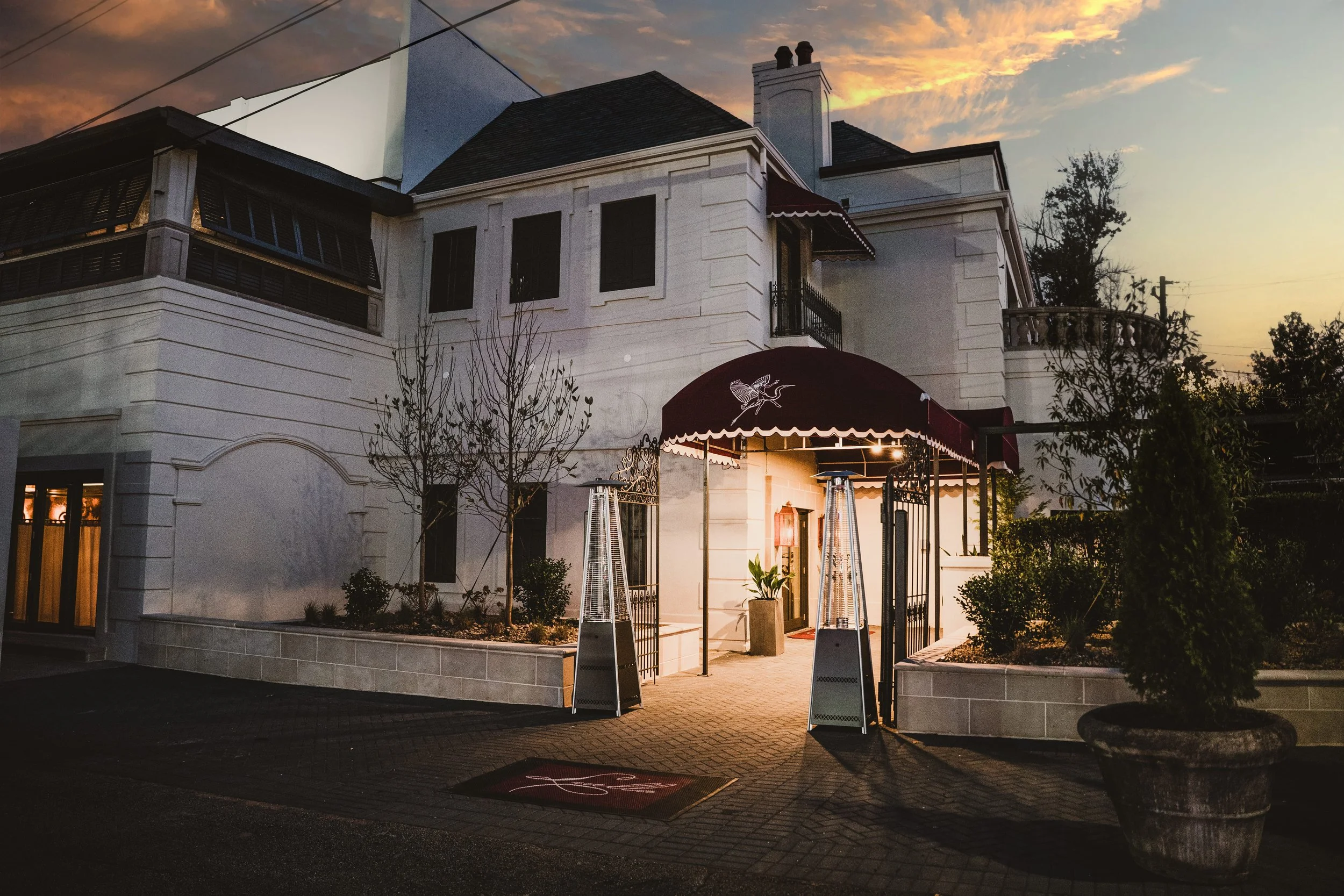 A white building with a maroon canopy over an entrance, illuminated at dusk. There are patio heaters, potted plants, leafless trees, and shrubbery around the entrance, with cars parked nearby.