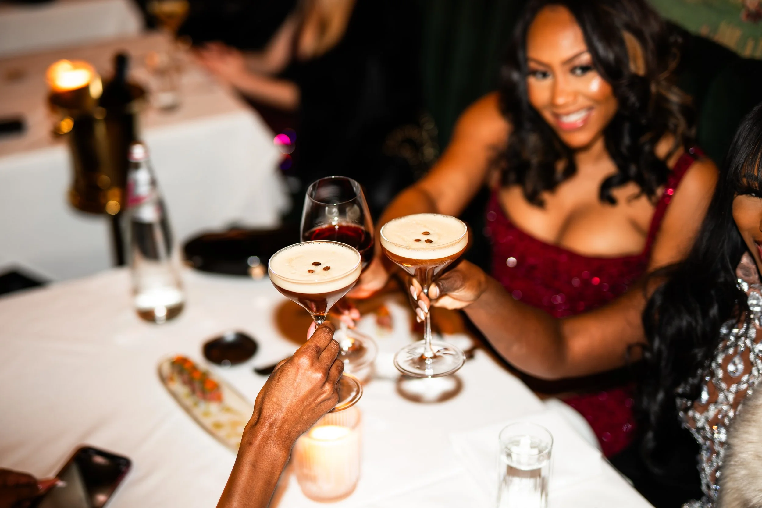 People celebrating and raising cocktails with a woman in a red dress smiling at a dinner table.