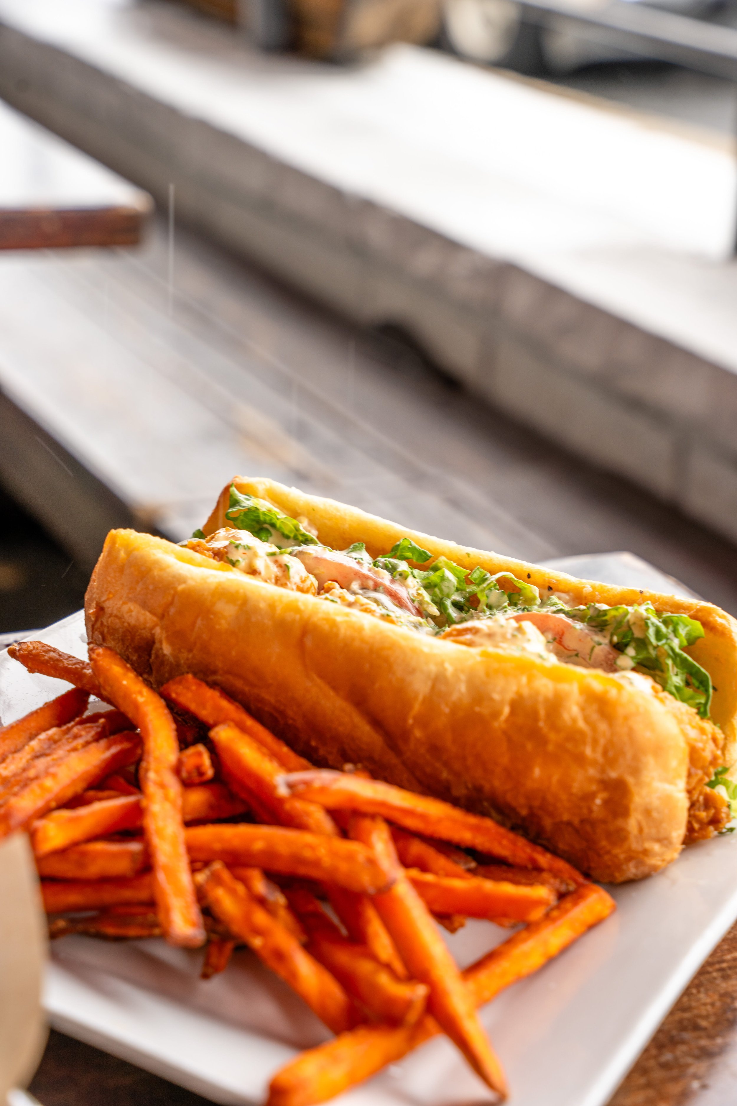 A plate with a fried fish po' boy sandwich filled with lettuce, tomatoes, and sauce, served with sweet potato fries.