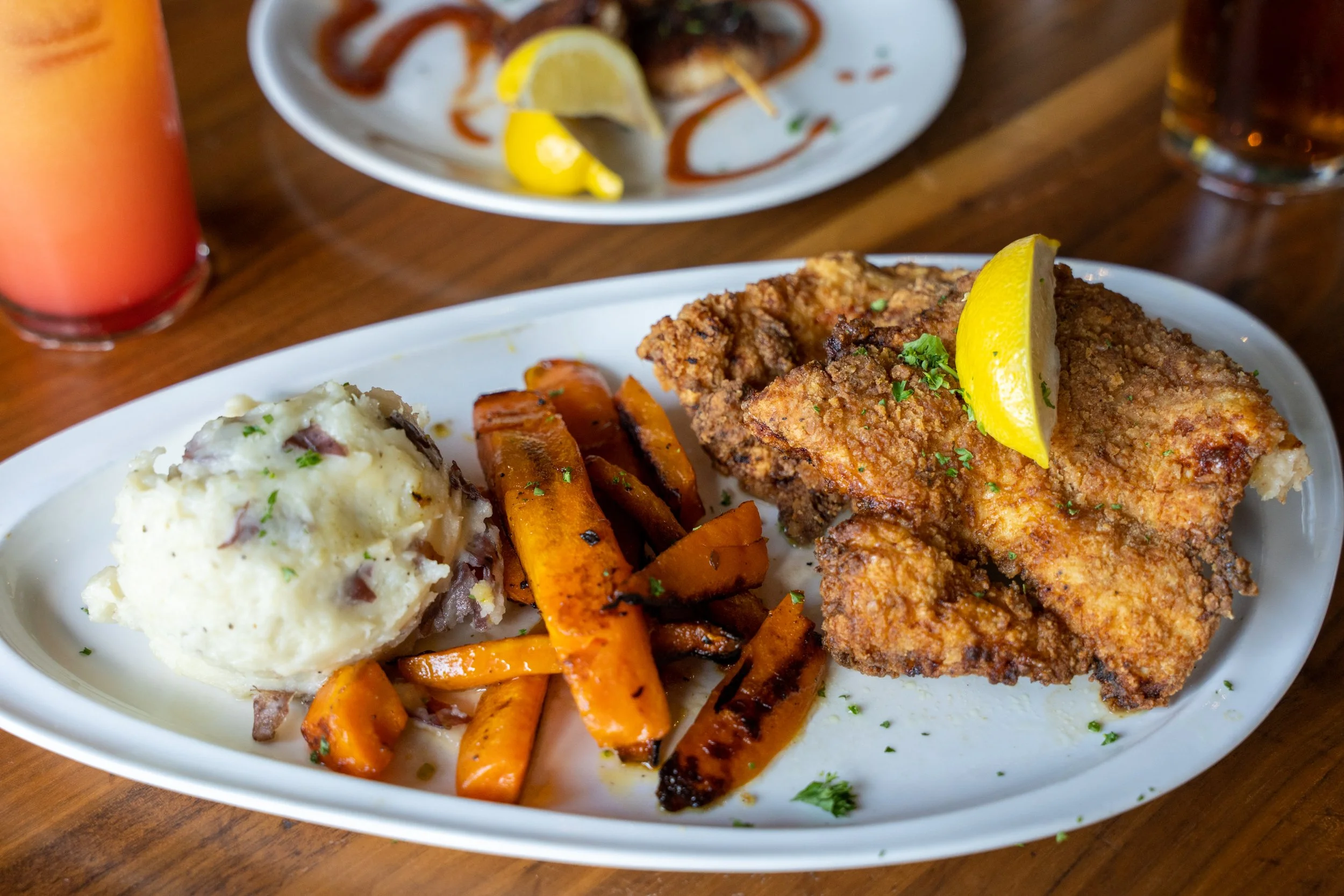 Fried fish fillet garnished with lemon wedge, mashed potatoes with herbs, and roasted carrots on a white oval plate.