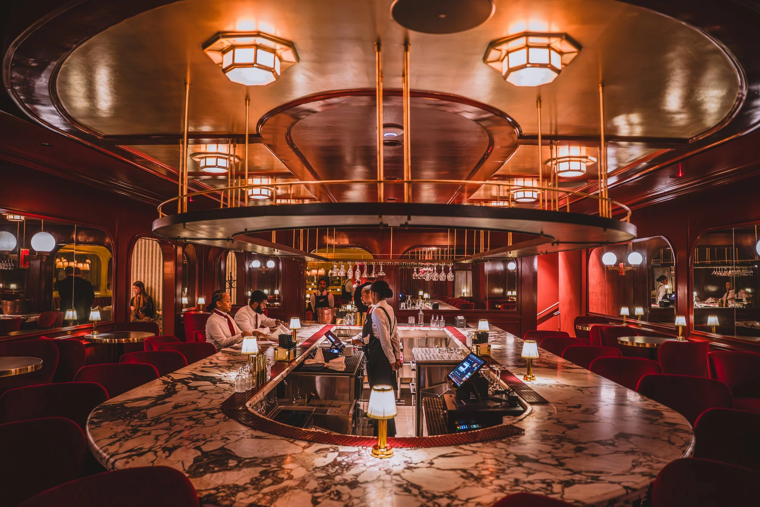 Interior of a luxurious bar with a marble counter, red velvet chairs, warm lighting, and staff preparing drinks.