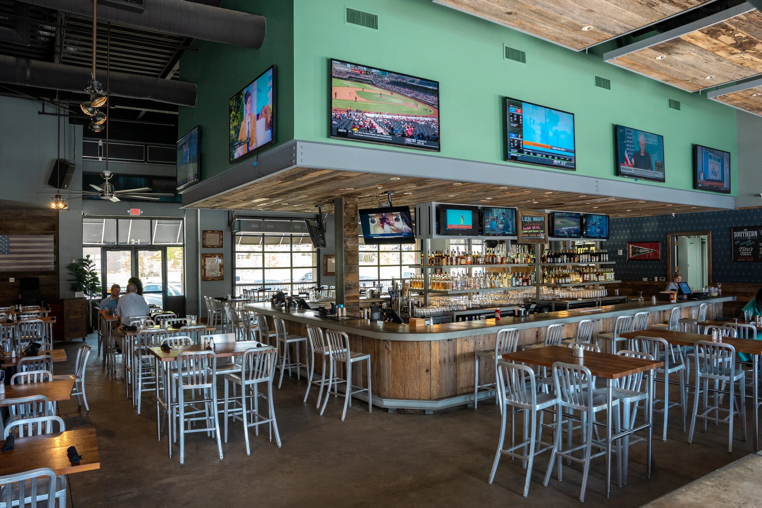 Empty bar and dining area inside a sports bar with multiple flat-screen TVs showing sports events, wooden tables and chairs, a bar with various bottles of alcohol, and windows letting in natural light.