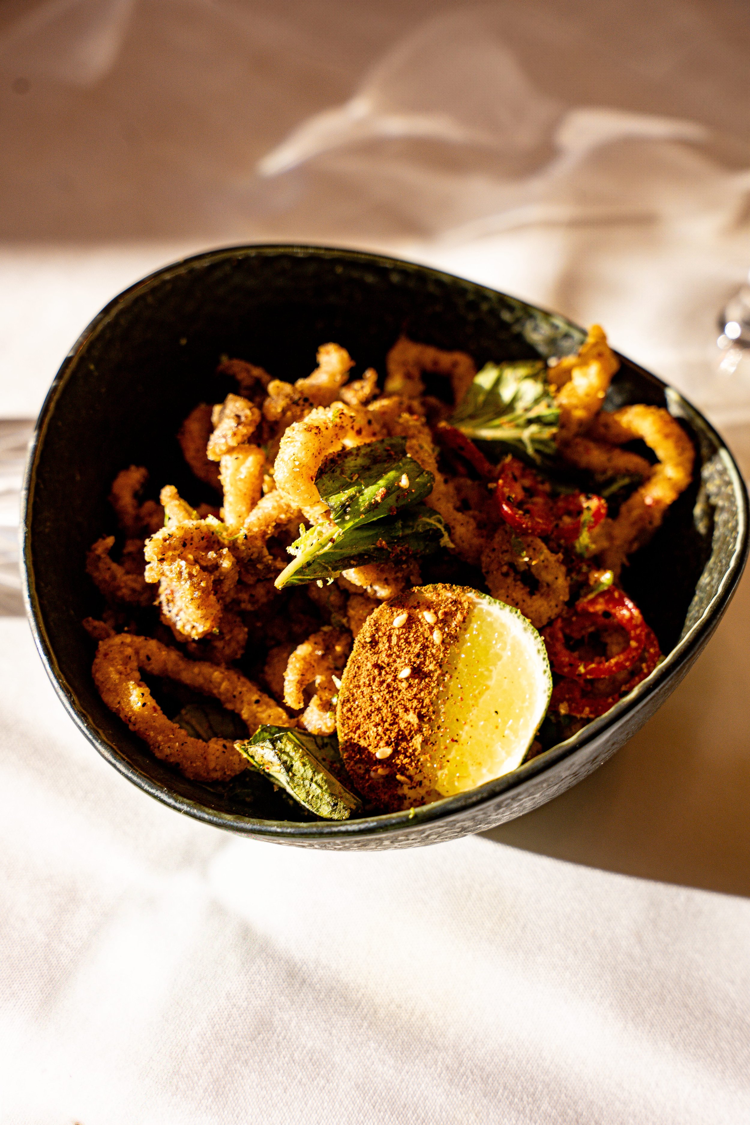 A black bowl filled with fried calamari garnished with basil leaves, a wedge of lemon, and red chili slices on a white tablecloth.