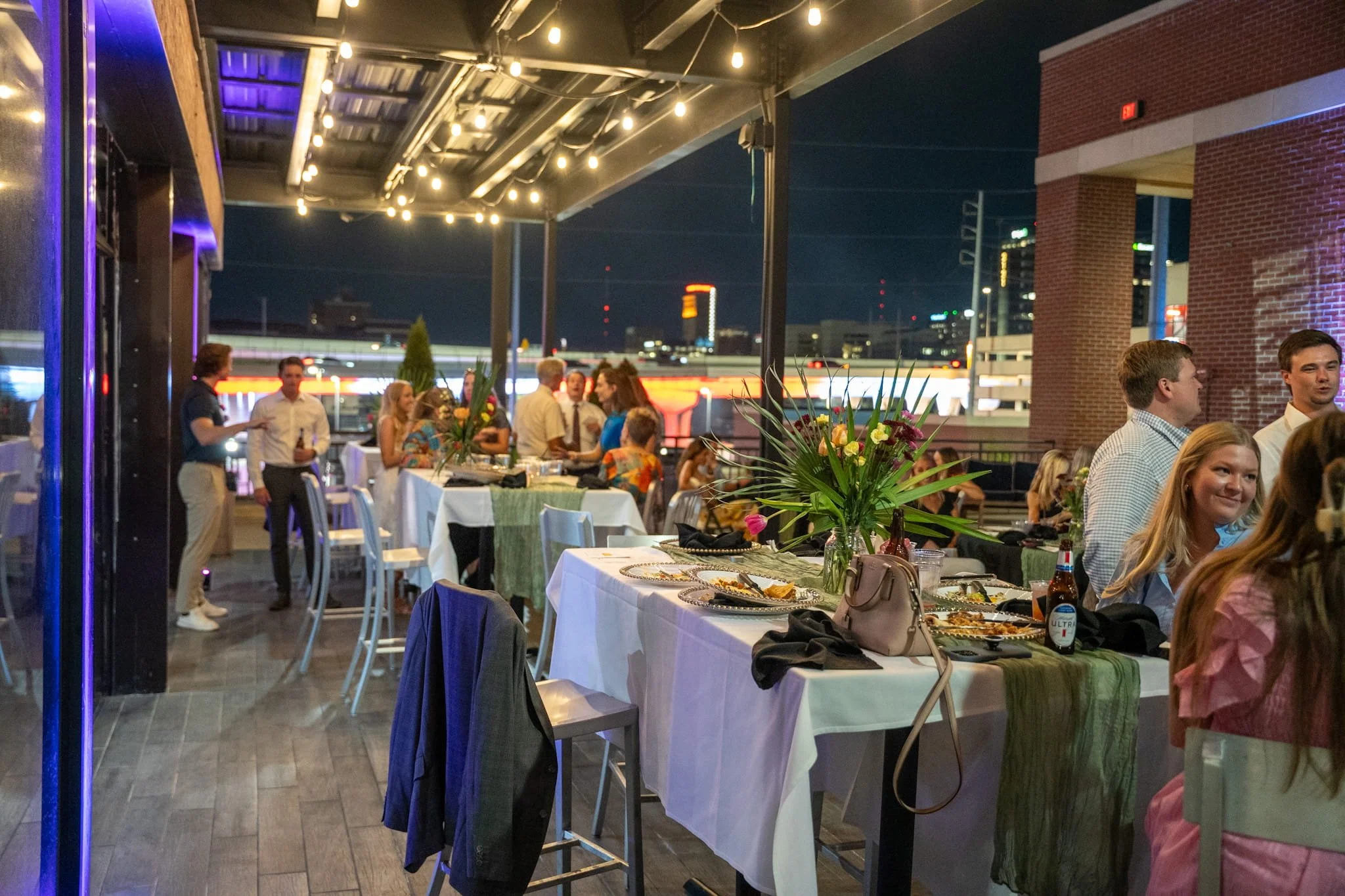 People gathered on an outdoor rooftop patio at night, celebrating with tables set with food, drinks, and floral centerpieces.