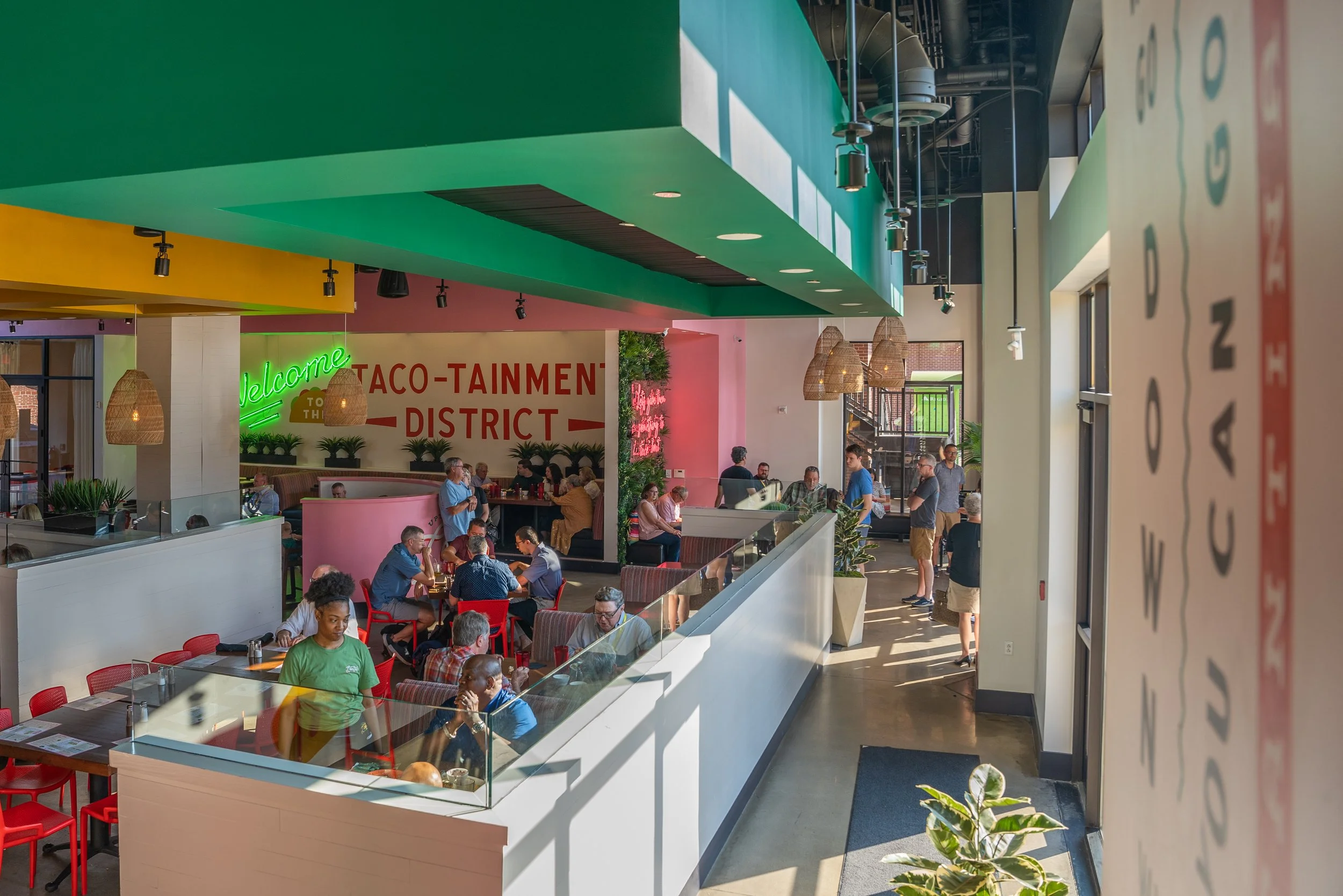 Interior of a modern restaurant or food court with colorful walls and ceiling, green, pink, and yellow accents, wooden pendant lights, and many people dining and waiting.