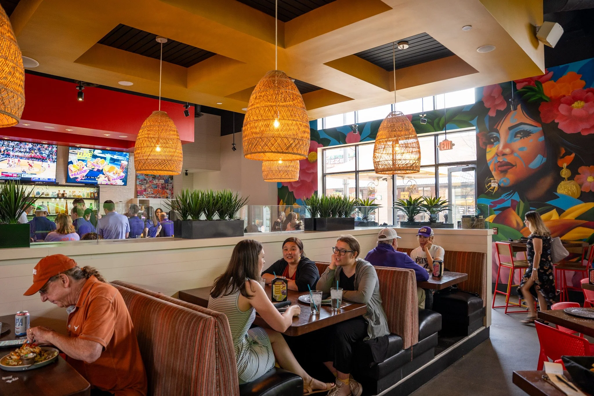 People dining and socializing inside a colorful restaurant with vibrant murals and hanging wicker lights.
