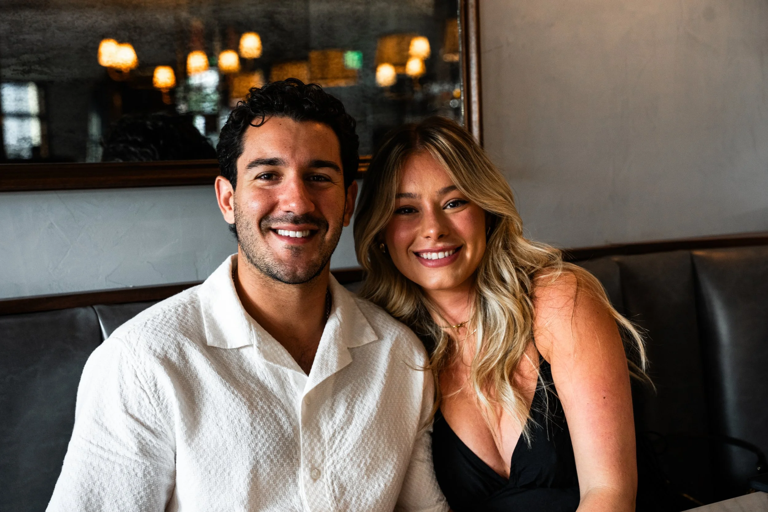 Smiling couple sitting together in a restaurant, with a dark leather booth and a mirror in the background.