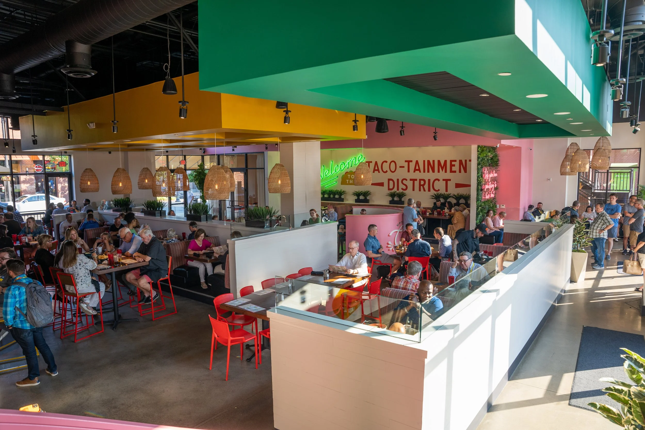 Interior of a bustling taco restaurant with colorful ceiling accents, hanging wicker lamps, and people dining and waiting in line.
