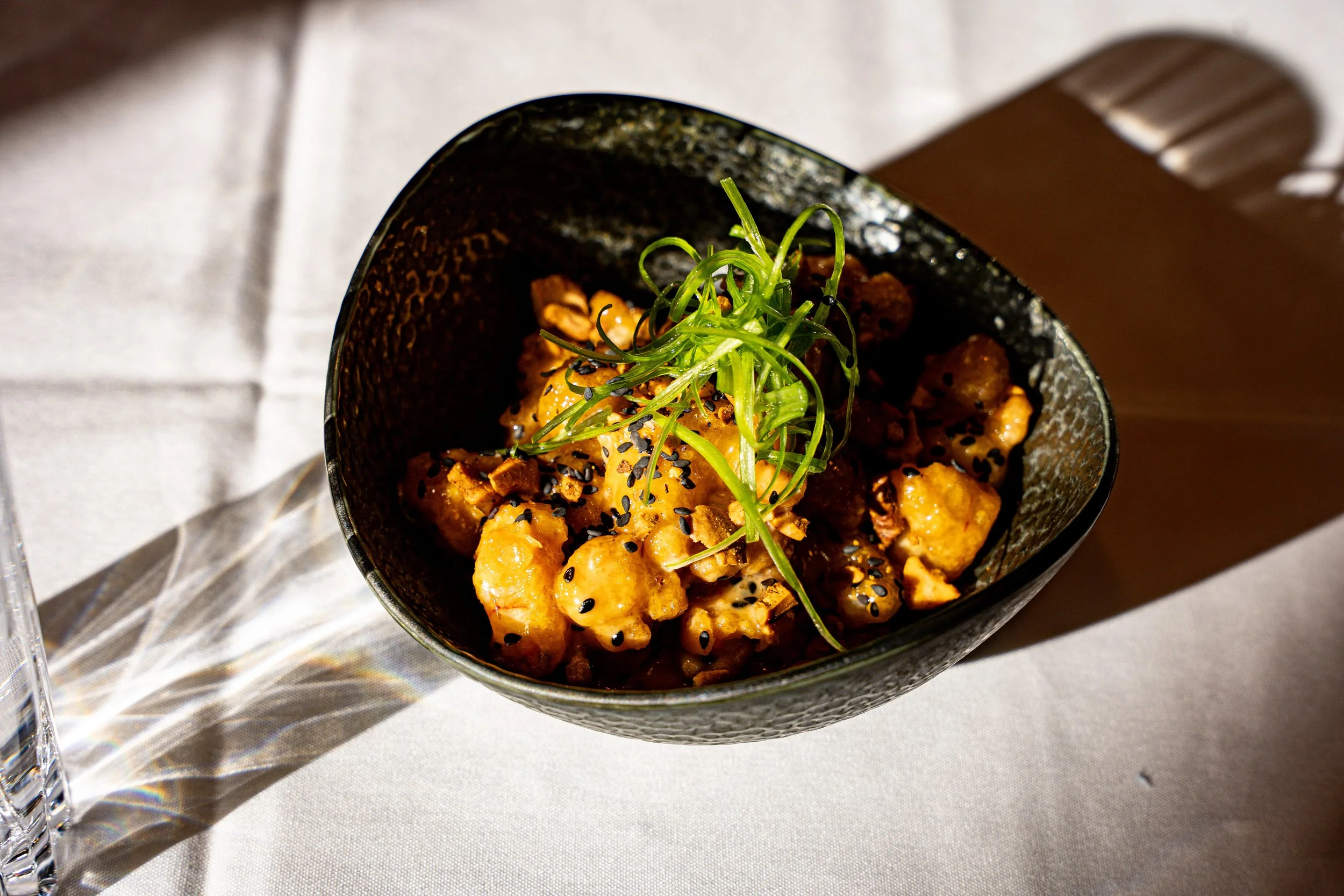 A black textured bowl containing glazed chicken pieces topped with chopped green onions and black sesame seeds, served on a white tablecloth.