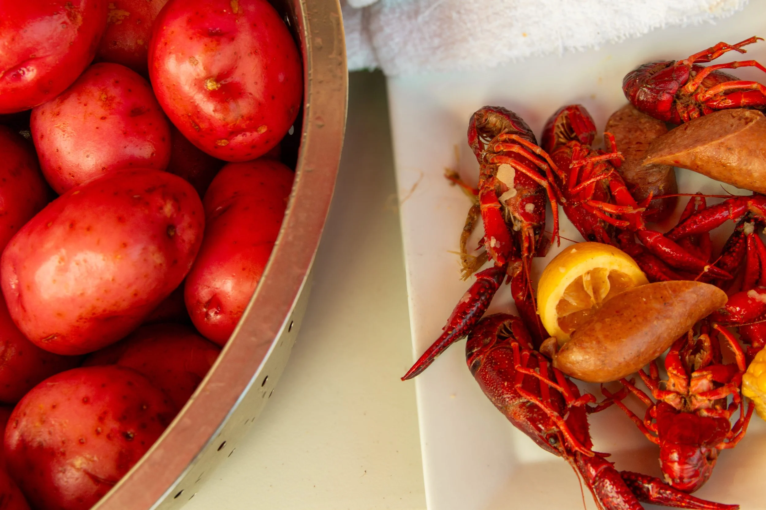 Fresh red potatoes in a metal basket on the left and cooked crawfish with lemon wedges and other seafood on a white platter on the right.