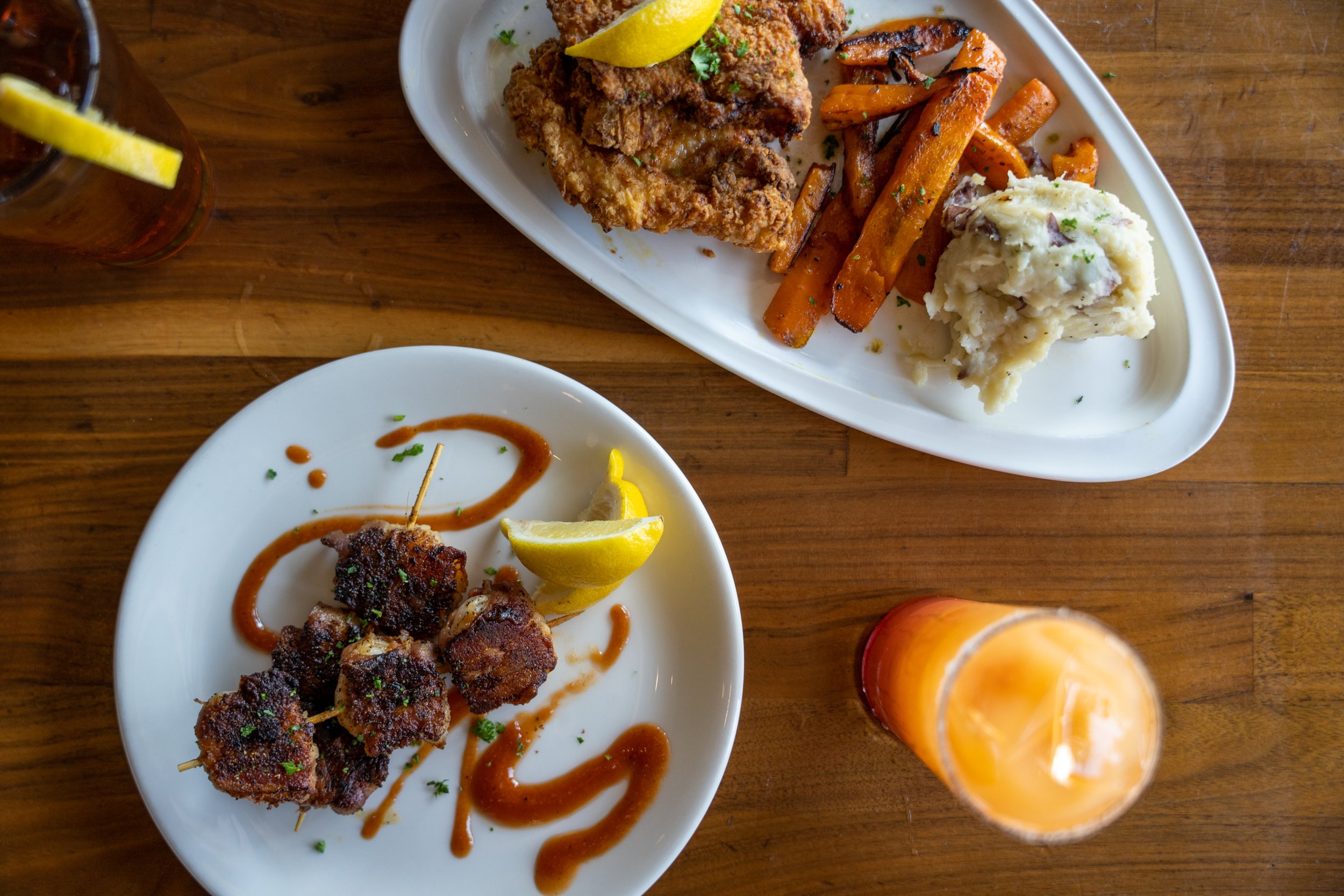 Two plates of food on a wooden table. The larger plate has fried chicken with a lemon wedge, roasted carrots with herbs, and mashed potatoes. The smaller plate has five pieces of glazed meat on a skewer with a lemon wedge and sauce drizzled around.