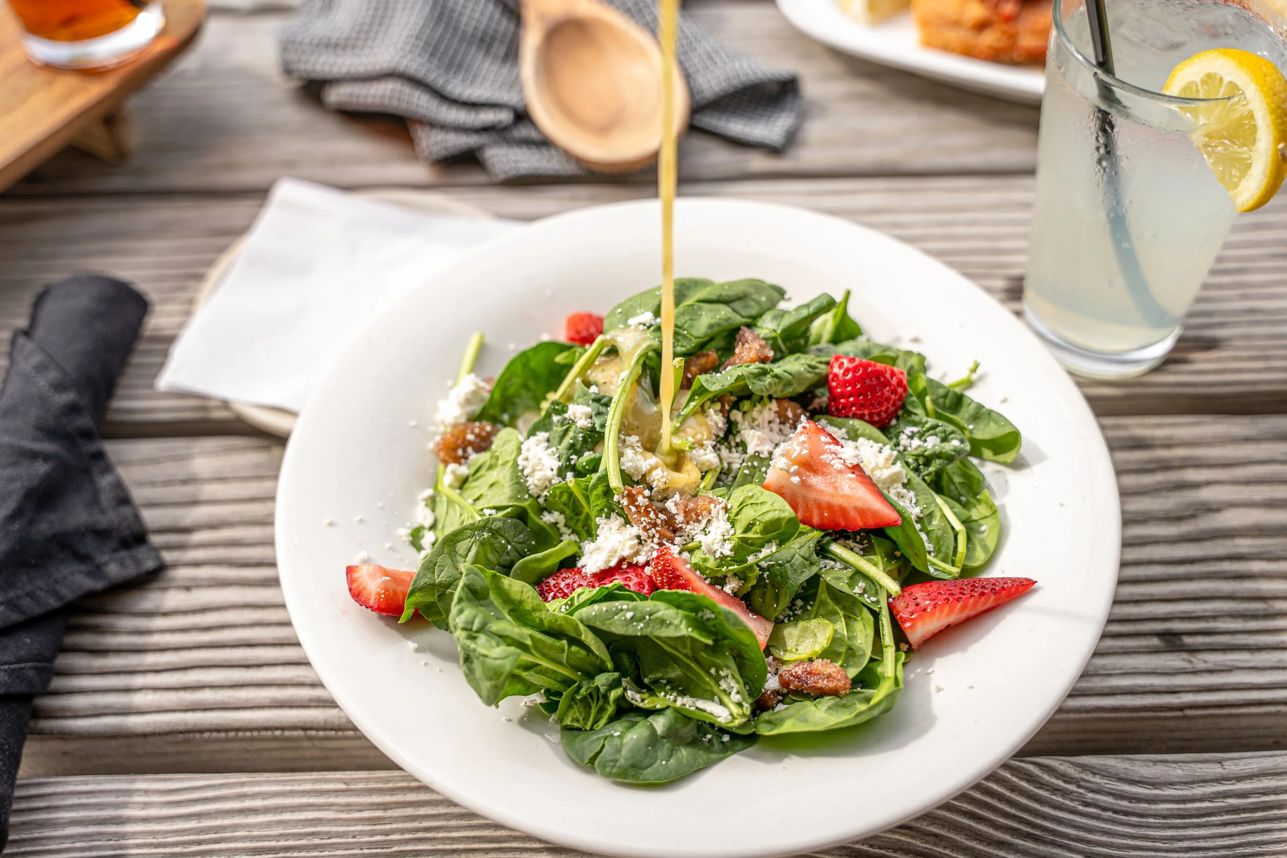 Fresh spinach salad with strawberries, cheese, and bacon, with dressing being poured on top, served on a white plate alongside a glass of lemonade with lemon slices, on a wooden table.
