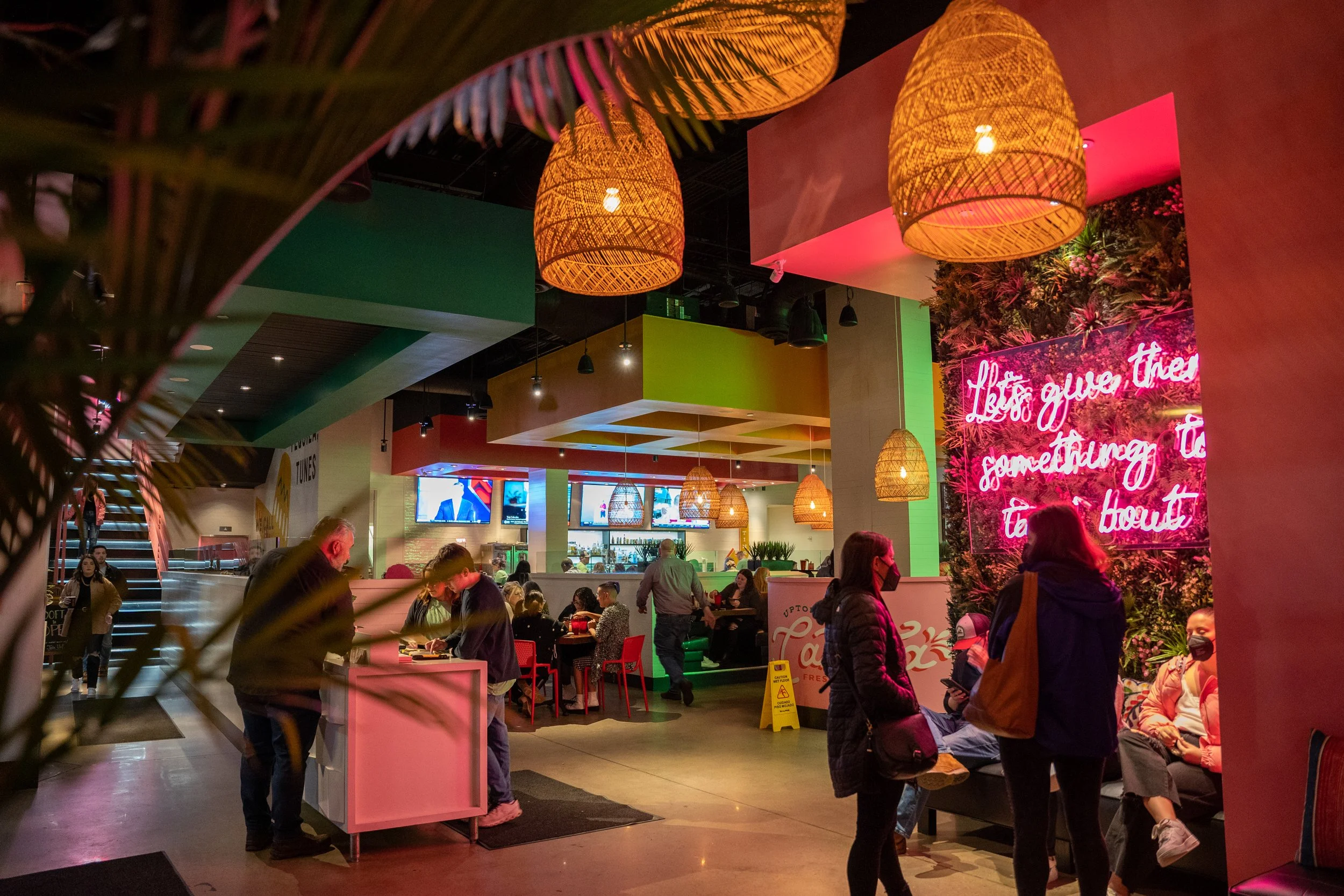 Interior of a lively restaurant with colorful decor, hanging wicker lamps, a neon pink sign that reads 'Let’s give these something to' and people sitting and standing in groups, some wearing masks.