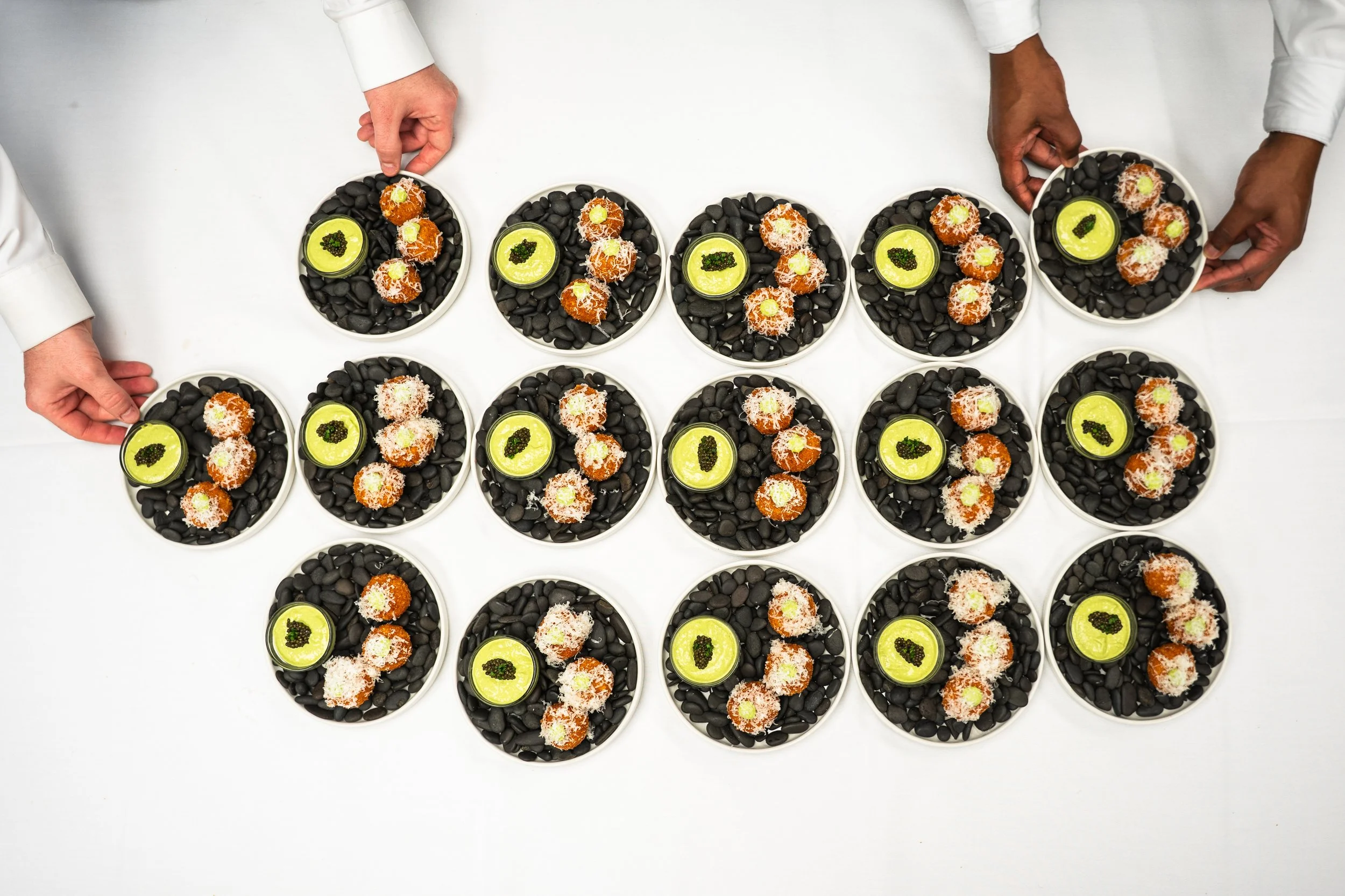 Multiple plates with black stones, each containing three small orange desserts topped with shredded coconut and a small green garnish, accompanied by a small round dish of green sauce with black garnish, on a white surface, with two people in white s
