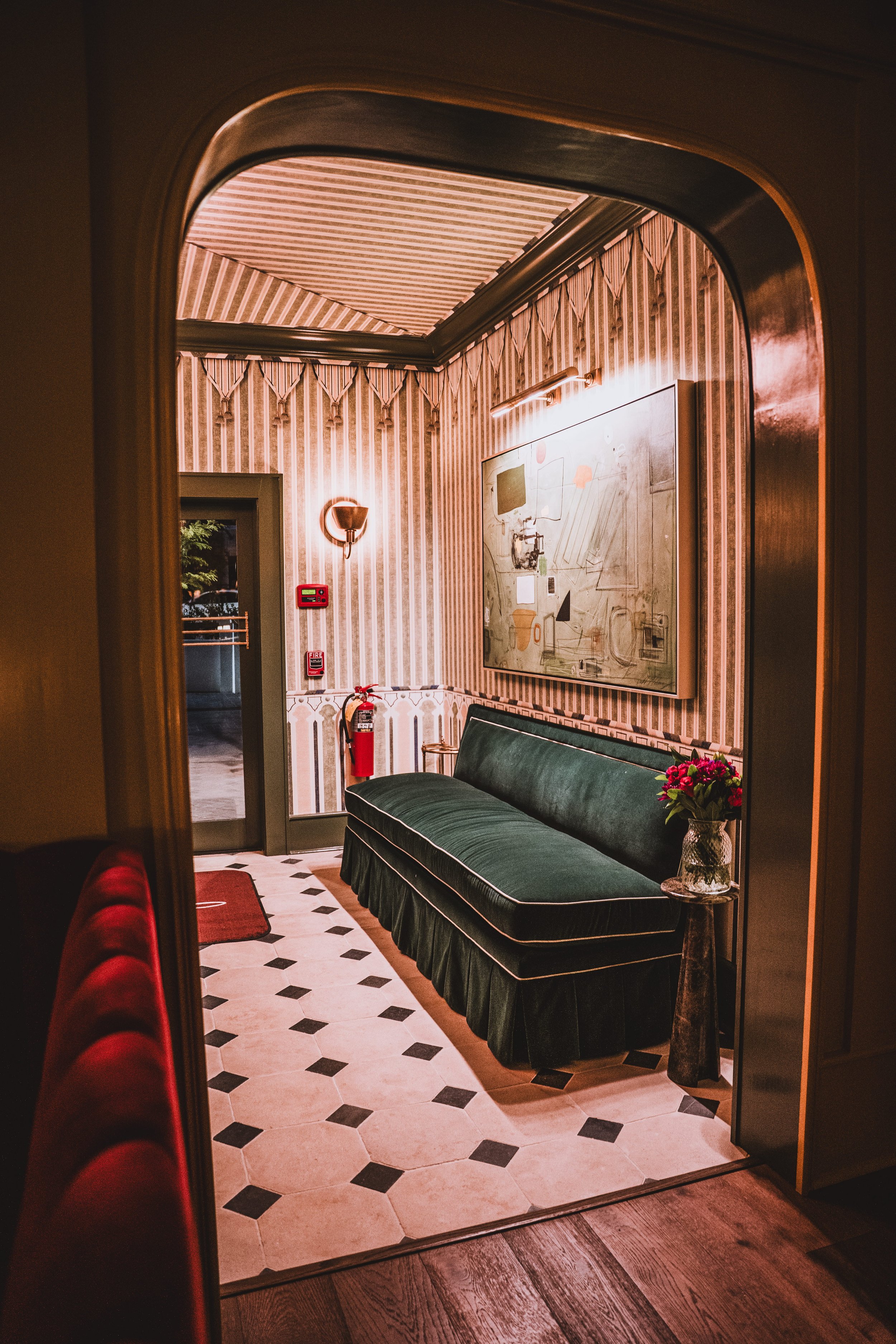 An interior view of a vintage-style waiting area with a green velvet bench, a small side table with a vase of pink flowers, a framed abstract artwork, and patterned wallpaper, illuminated by warm lighting.