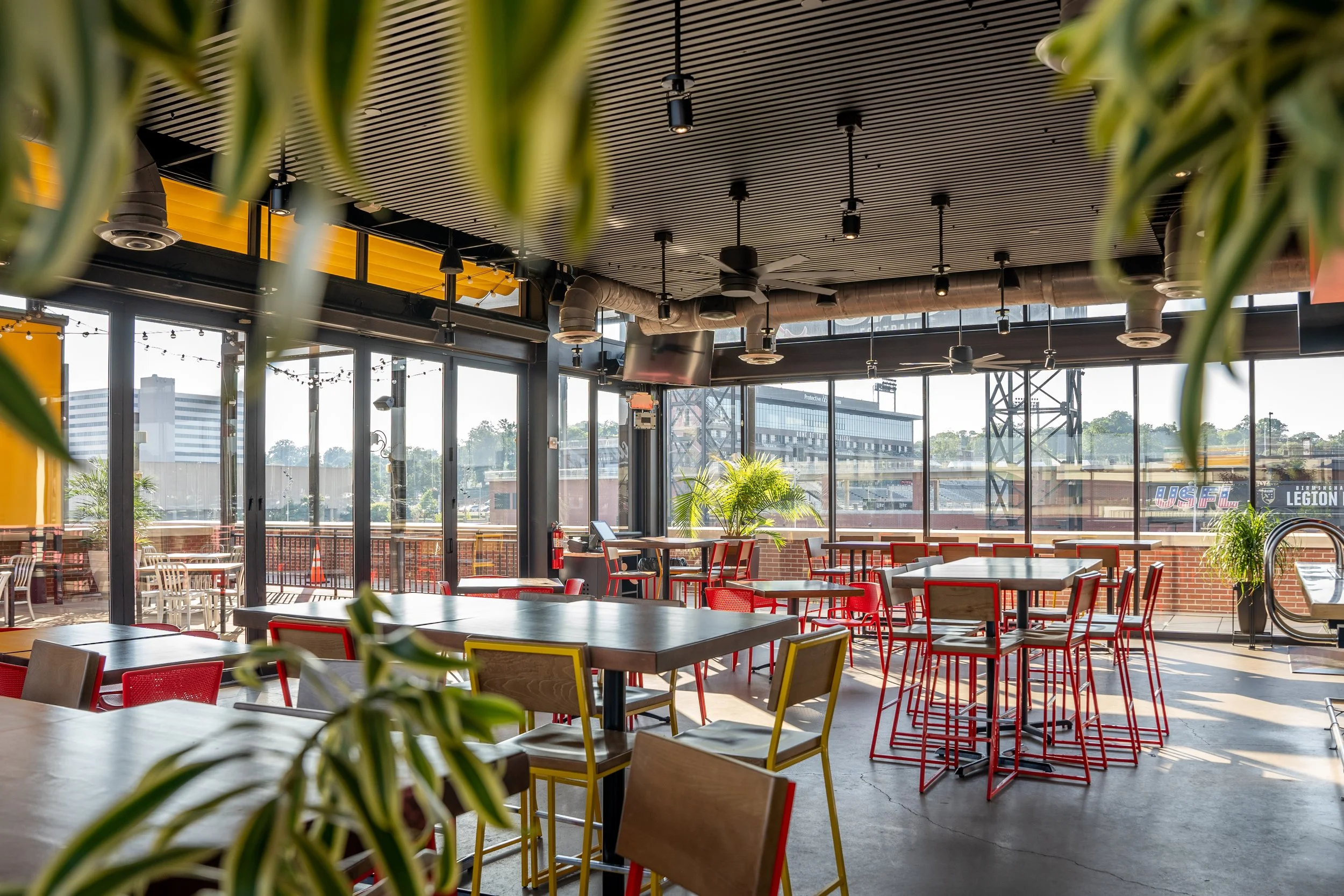 Interior of a modern, well-lit restaurant or cafe with large windows, red and yellow chairs, potted plants, and ceiling fans.
