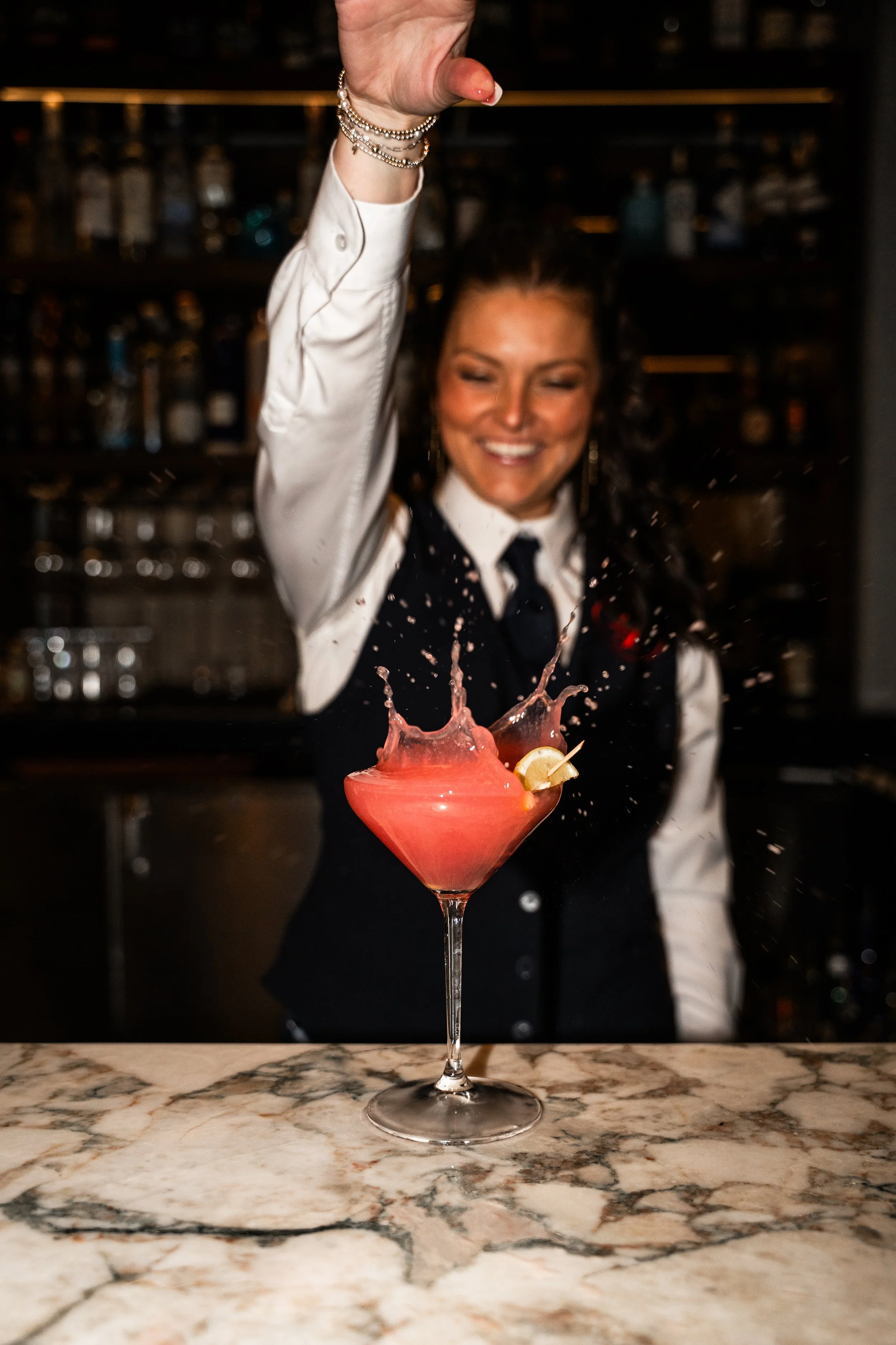 A bartender in a black vest and white shirt throws a pink cocktail into the air, causing a splash, while smiling behind a marble bar counter.
