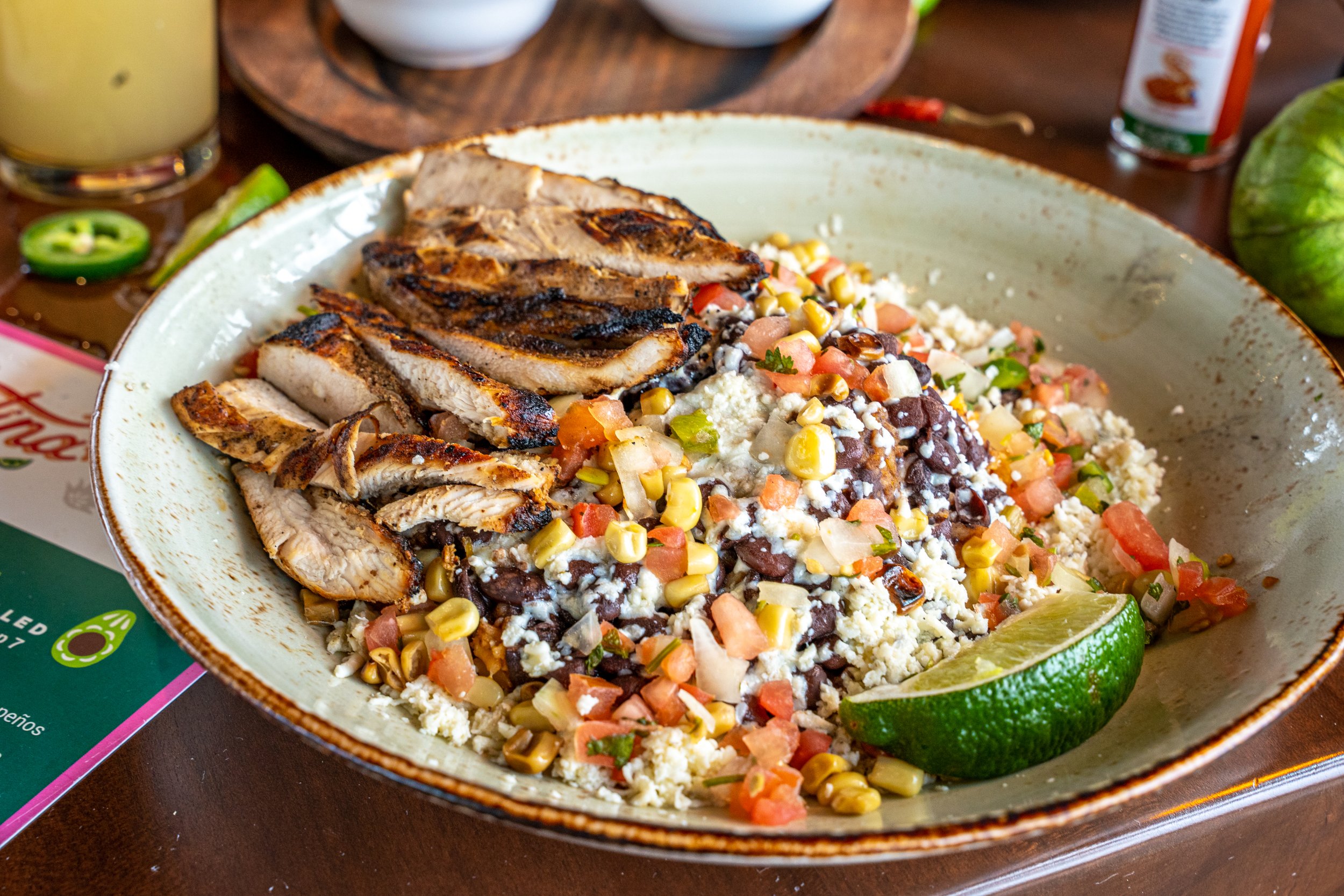 A bowl of Mexican-style street corn salad with grilled chicken slices and a lime wedge, garnished with vegetables and cheese, on a wooden table.