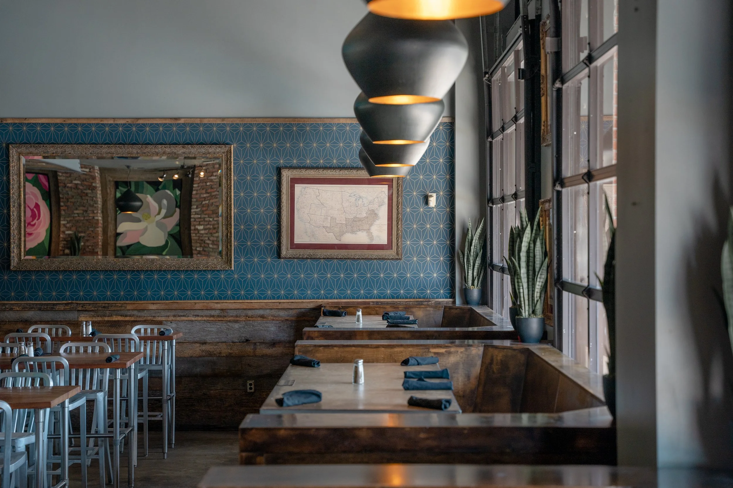 Empty restaurant with wooden tables set with napkins and salt shakers, blue patterned wallpaper, potted plants near large windows, framed map on wall, large mirror reflecting part of area, hanging pendant lights above tables.